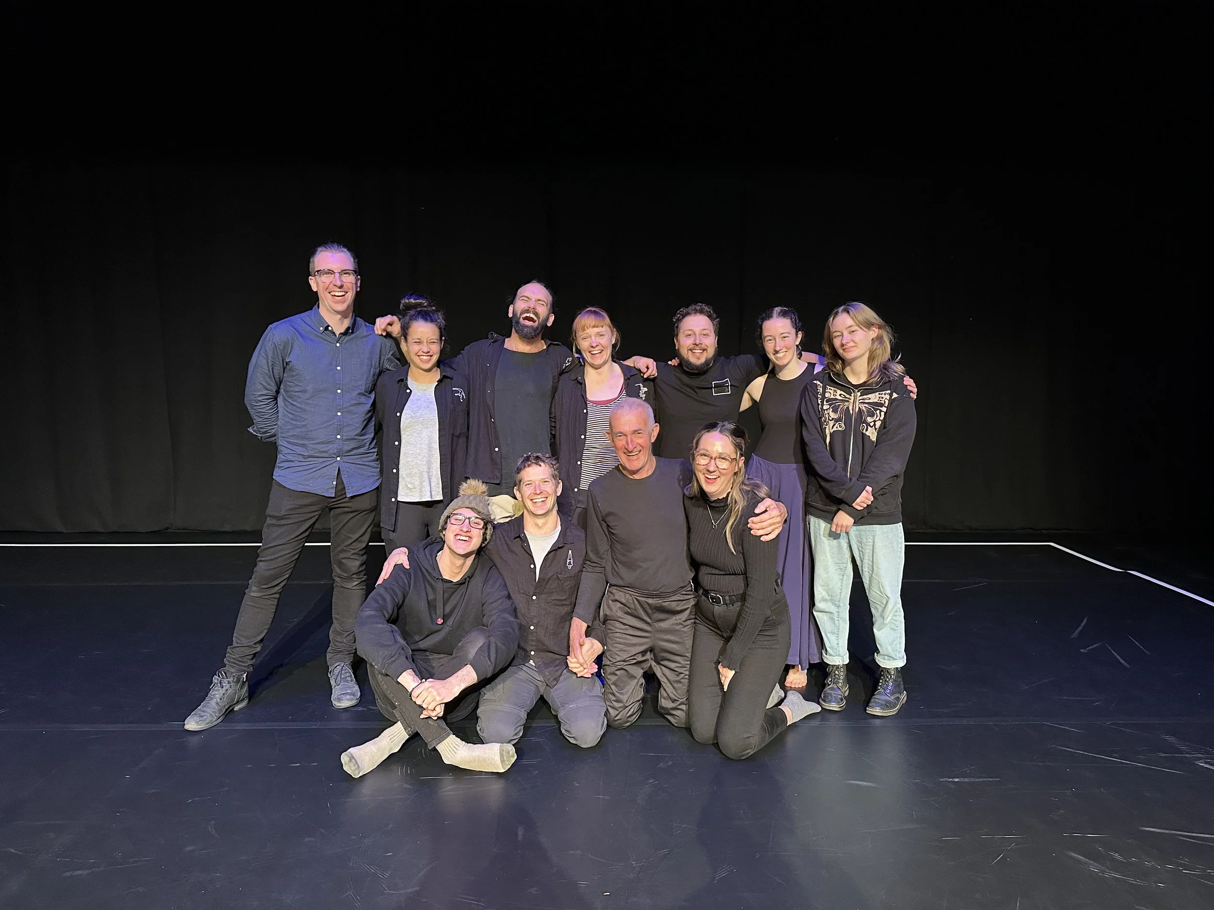 A group of eleven diverse people, smiling and posing together, on a black stage with a plain black background.