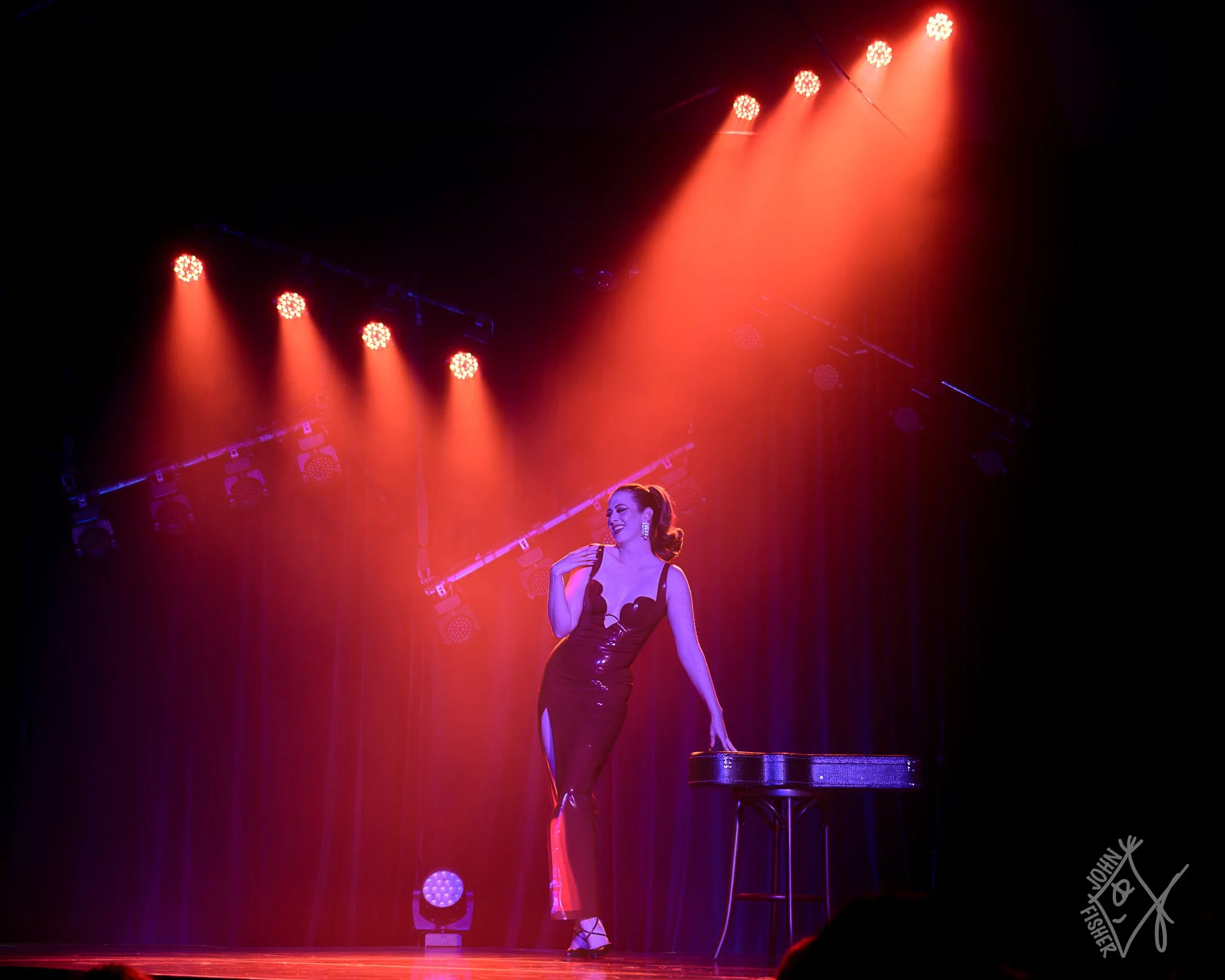 A woman lit in blue wearing a red latex dress leaning on a guitar case