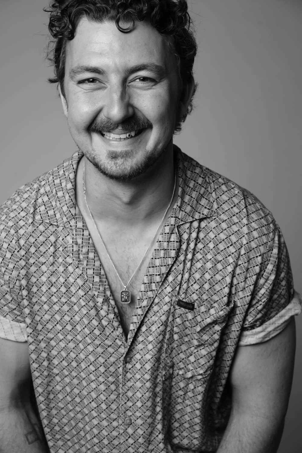 Black-and-white portrait of a smiling man with curly hair and a mustache, wearing a patterned shirt and a necklace.
