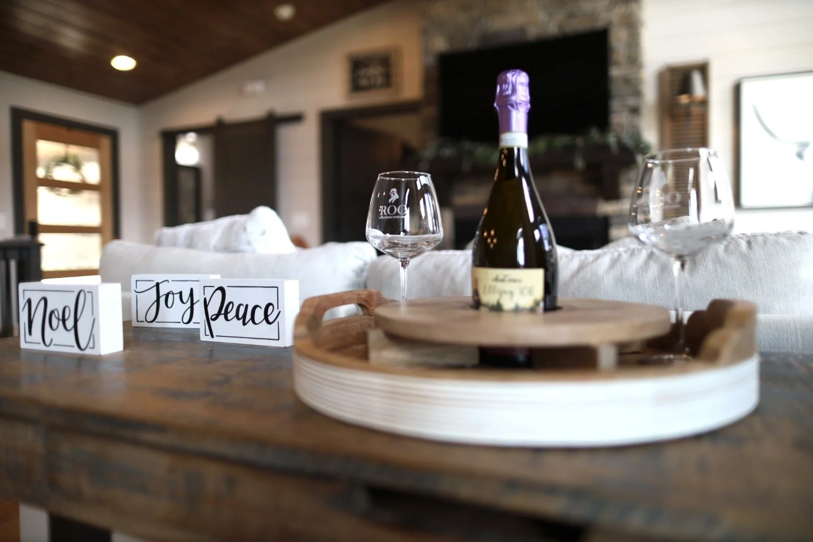 A festive table set with a bottle of wine, two wine glasses, and holiday signs that read 'Noel,' 'Joy,' and 'Peace.' The background shows a cozy living room with a white couch, a TV, a fireplace, and warm lighting.