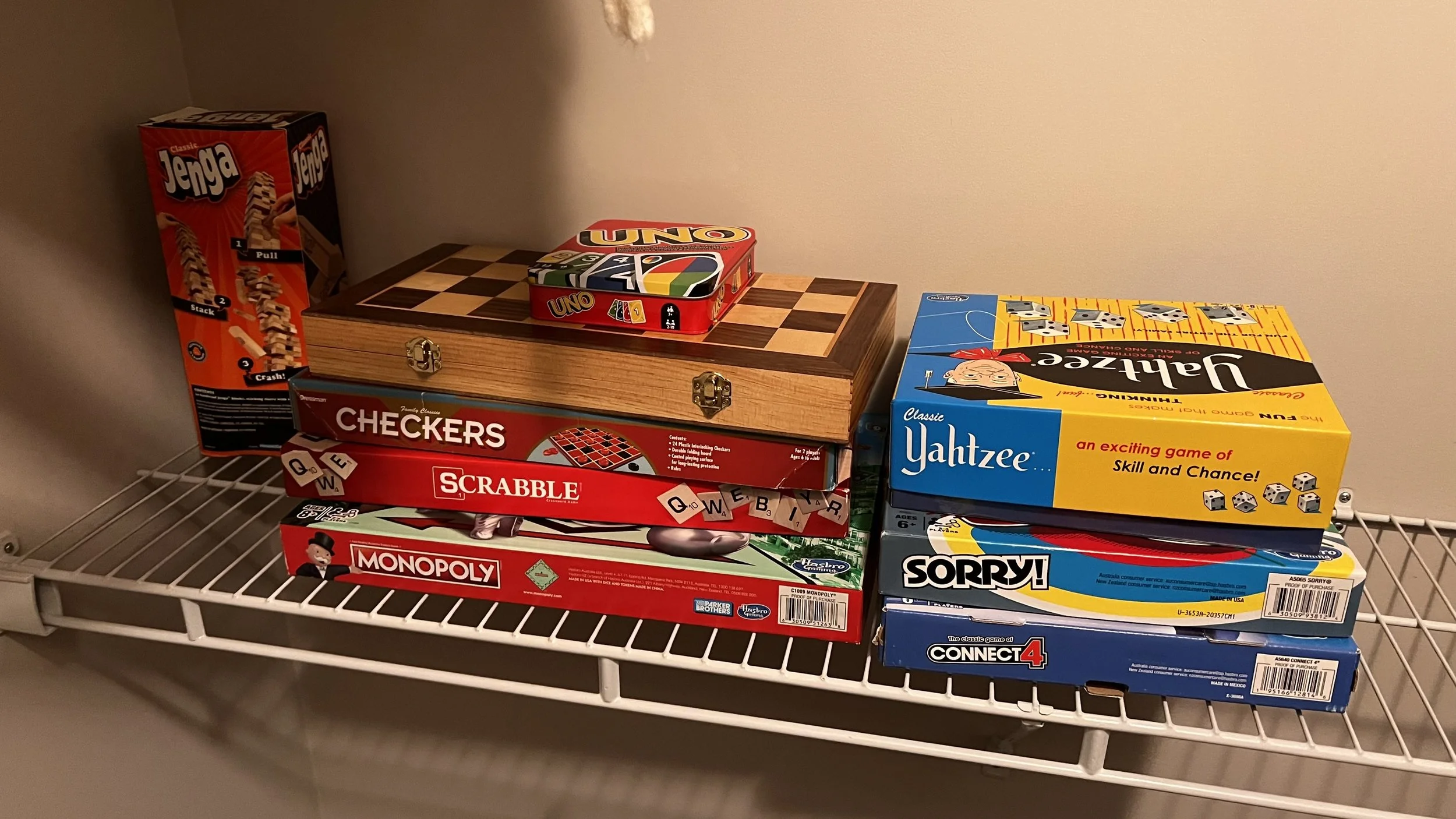Stacked board games on a metal shelf, including Jenga, Checkers, Scrabble, Monopoly, Yahtzee, Sorry!, and Connect 4, with a chessboard-patterned box on top.