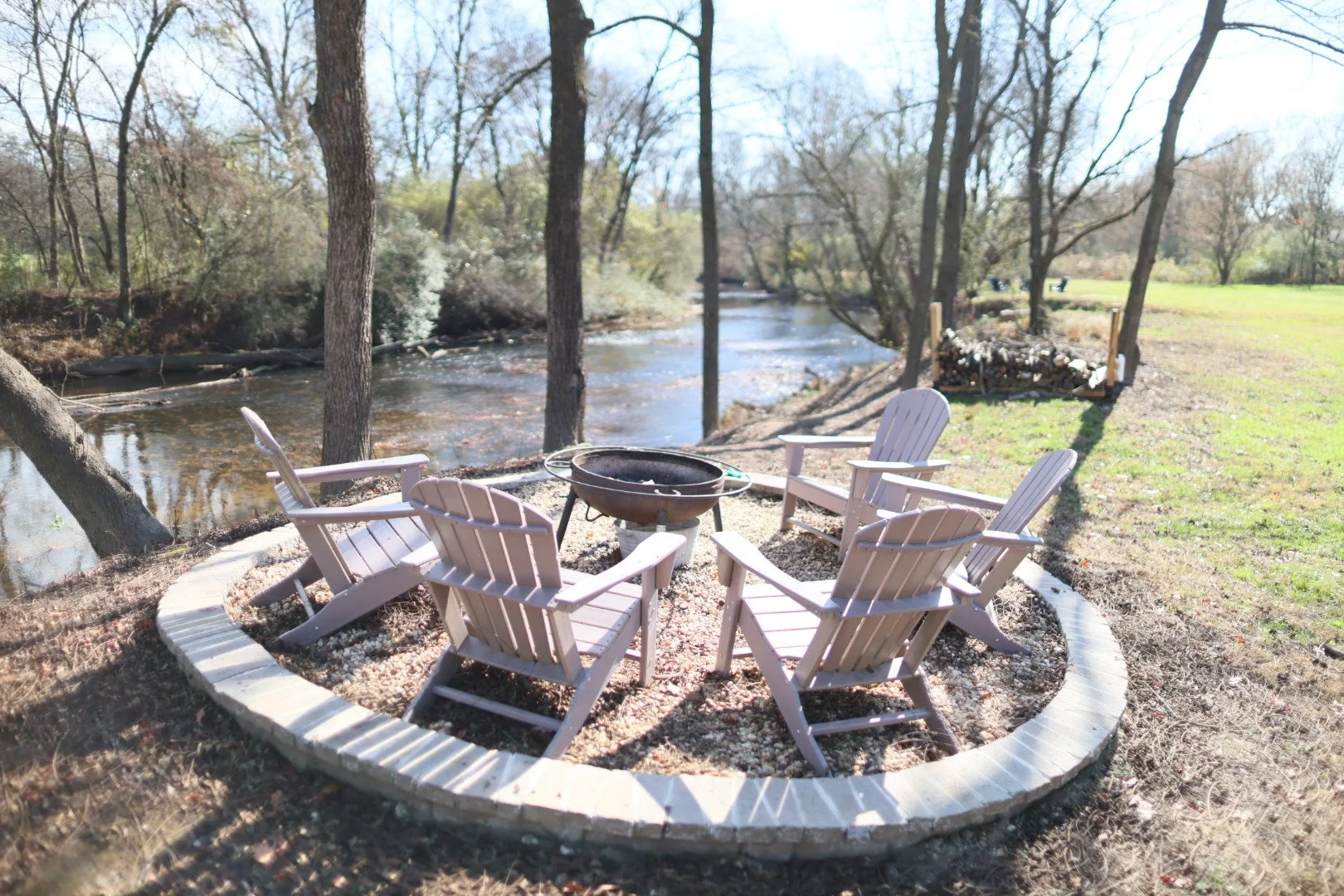 A circular seating area with six white Adirondack chairs arranged around a fire pit, located near a river with trees and greenery in the background.
