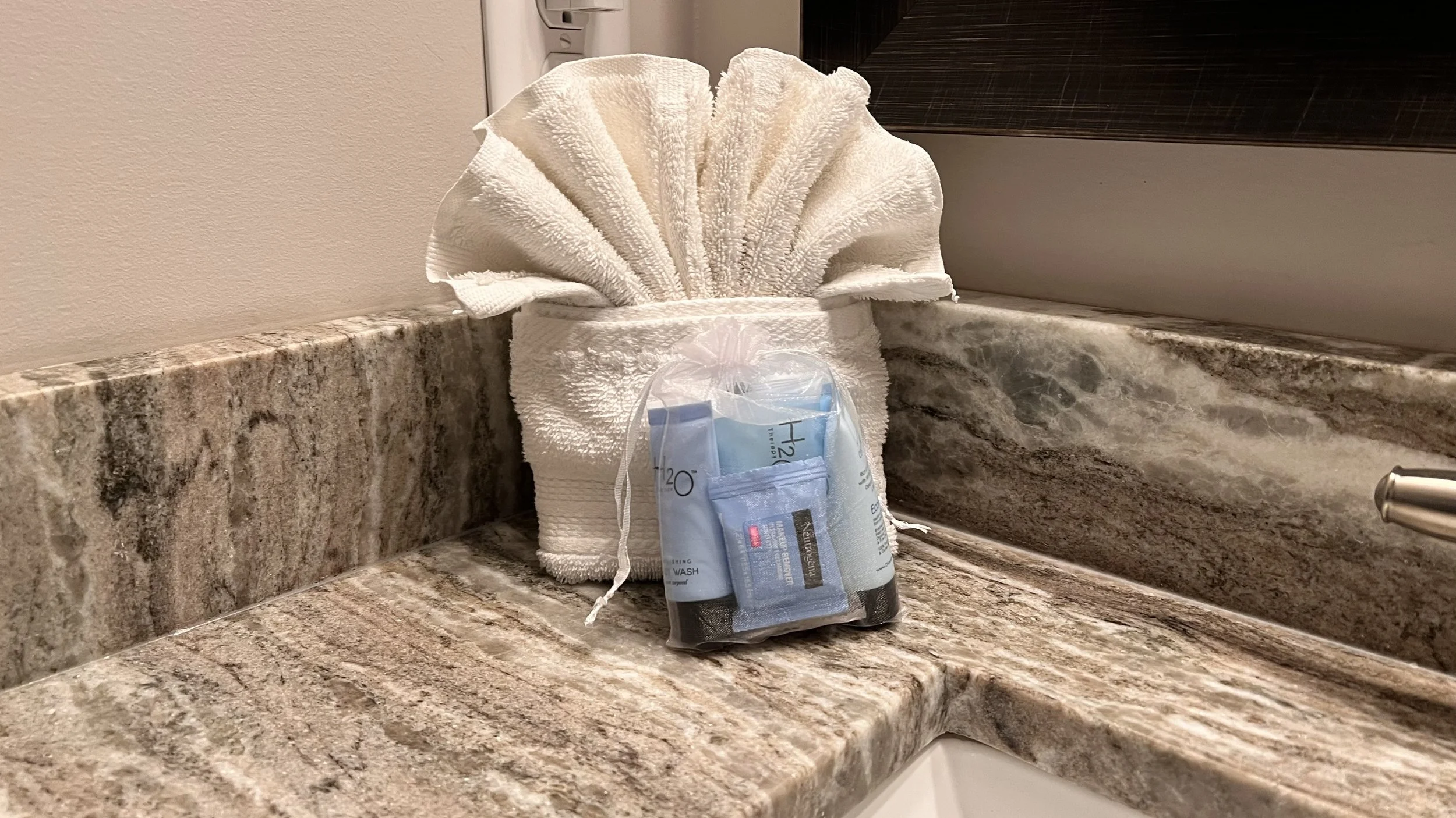 Towel folded into a fan shape on a marble countertop with small toiletry bottles in a plastic bag in front of it.