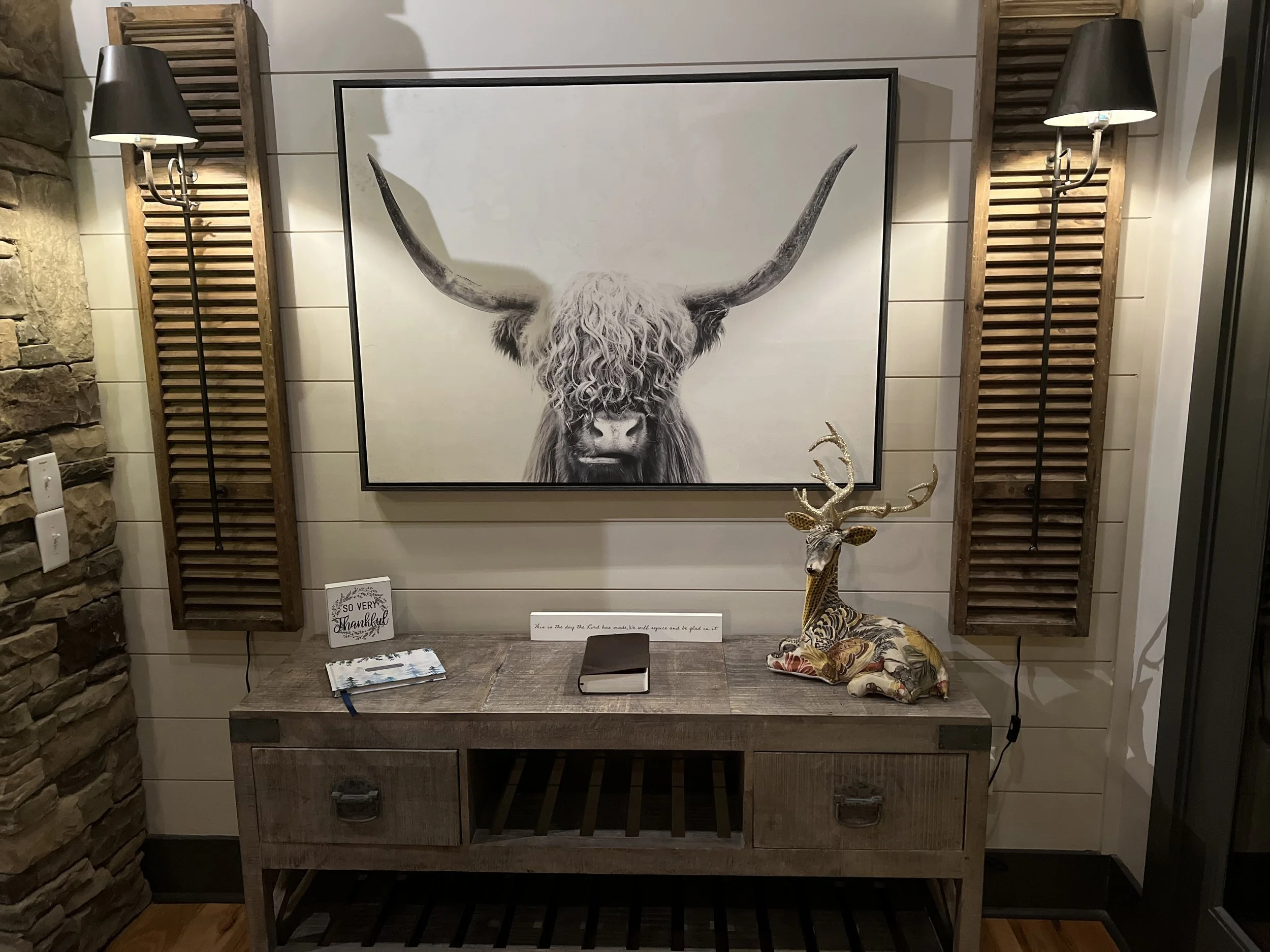 Wall decor with a large black and white painting of a Highland cow with long horns, flanked by wooden shutters with black lampshades. A wooden console table below holds a small sign, a closed book, and a decorative fabric-covered reindeer sculpture.
