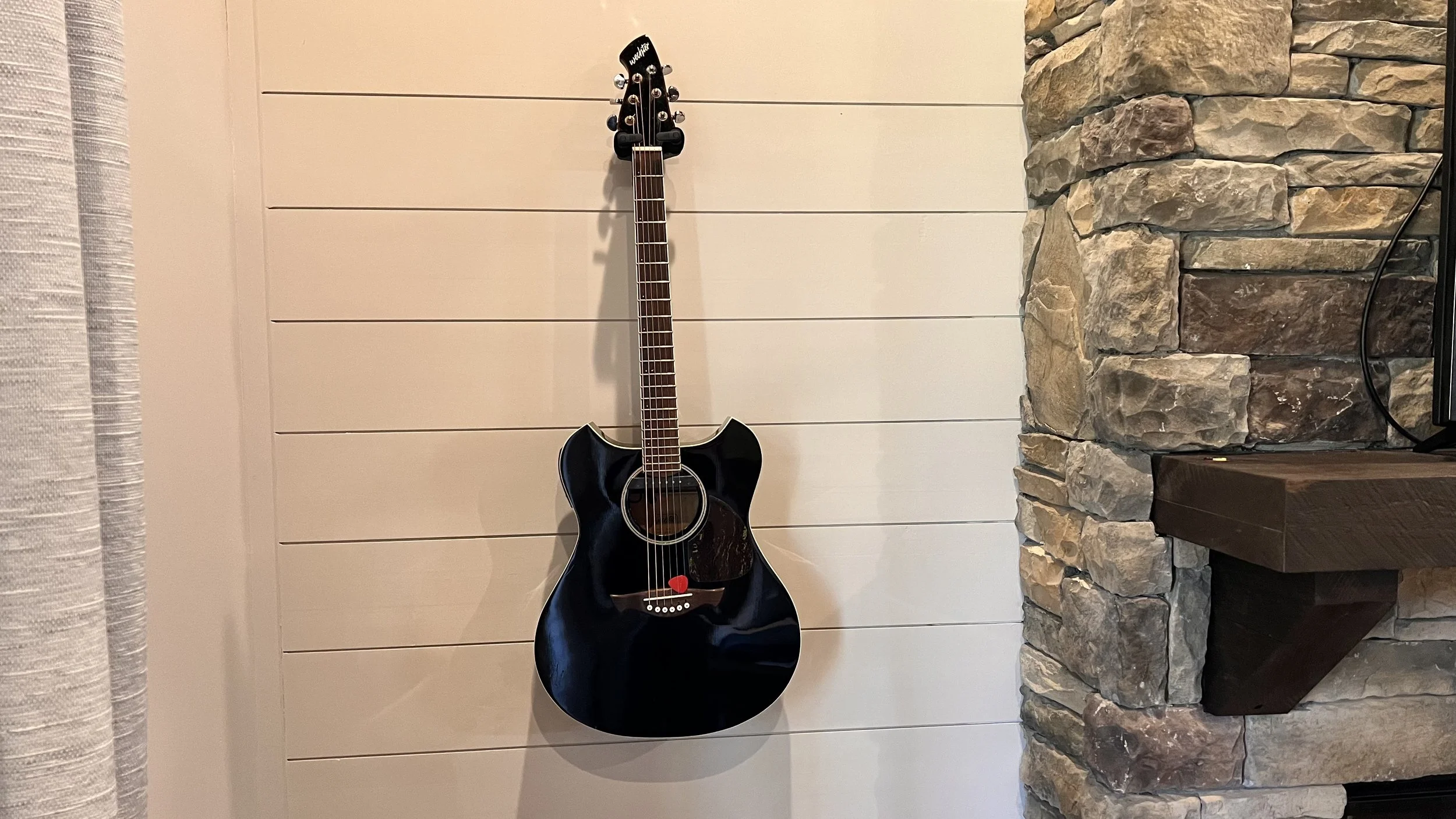 Black acoustic guitar hanging on a white wooden paneled wall beside a stone fireplace.