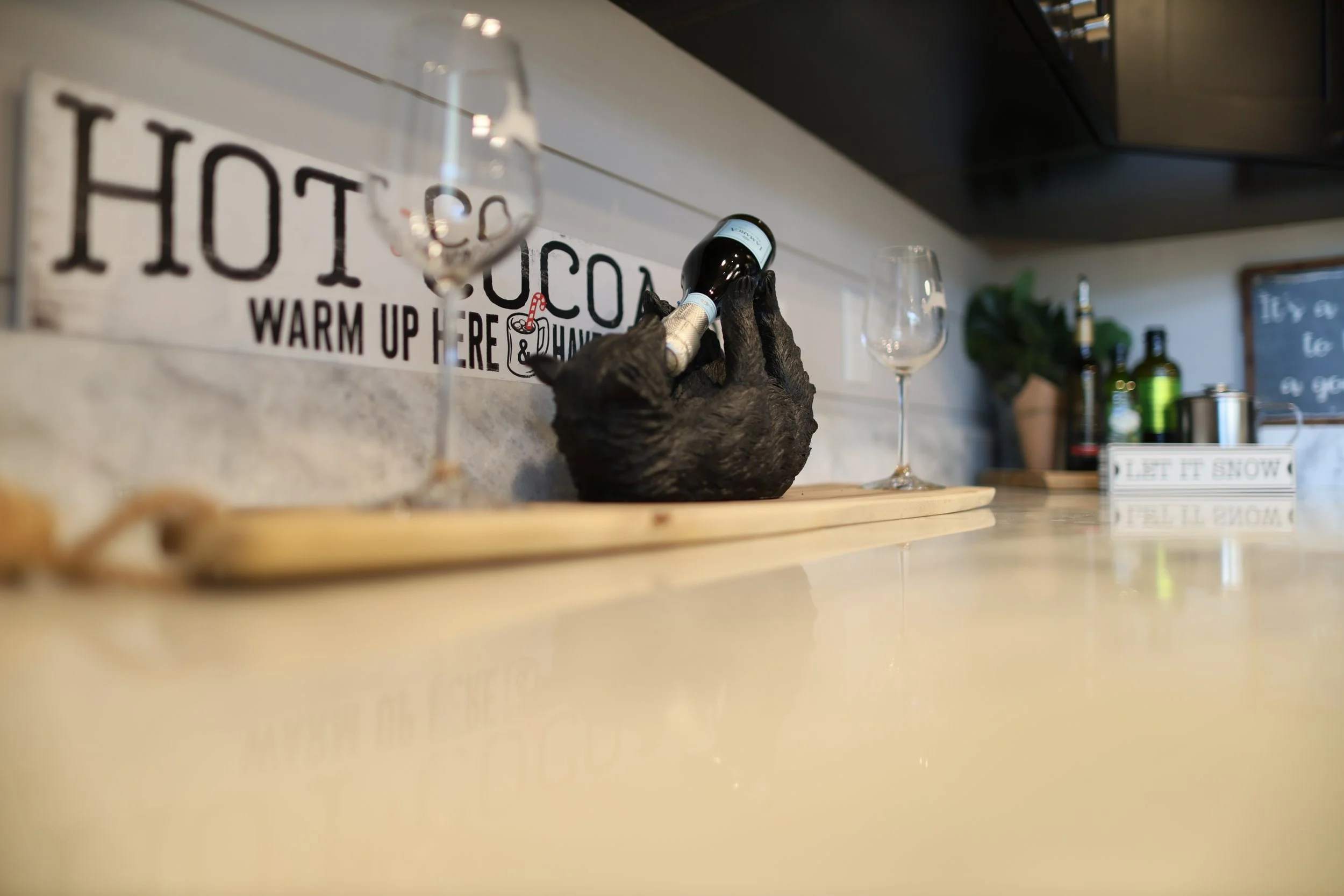A bar countertop with a black bear shaped holder holding a small wine bottle, two empty wine glasses, bottles of alcohol, a small chalkboard, and a decorative box with the text "Let It Snow." In the background is a wall sign that reads "Hot Cocoa War