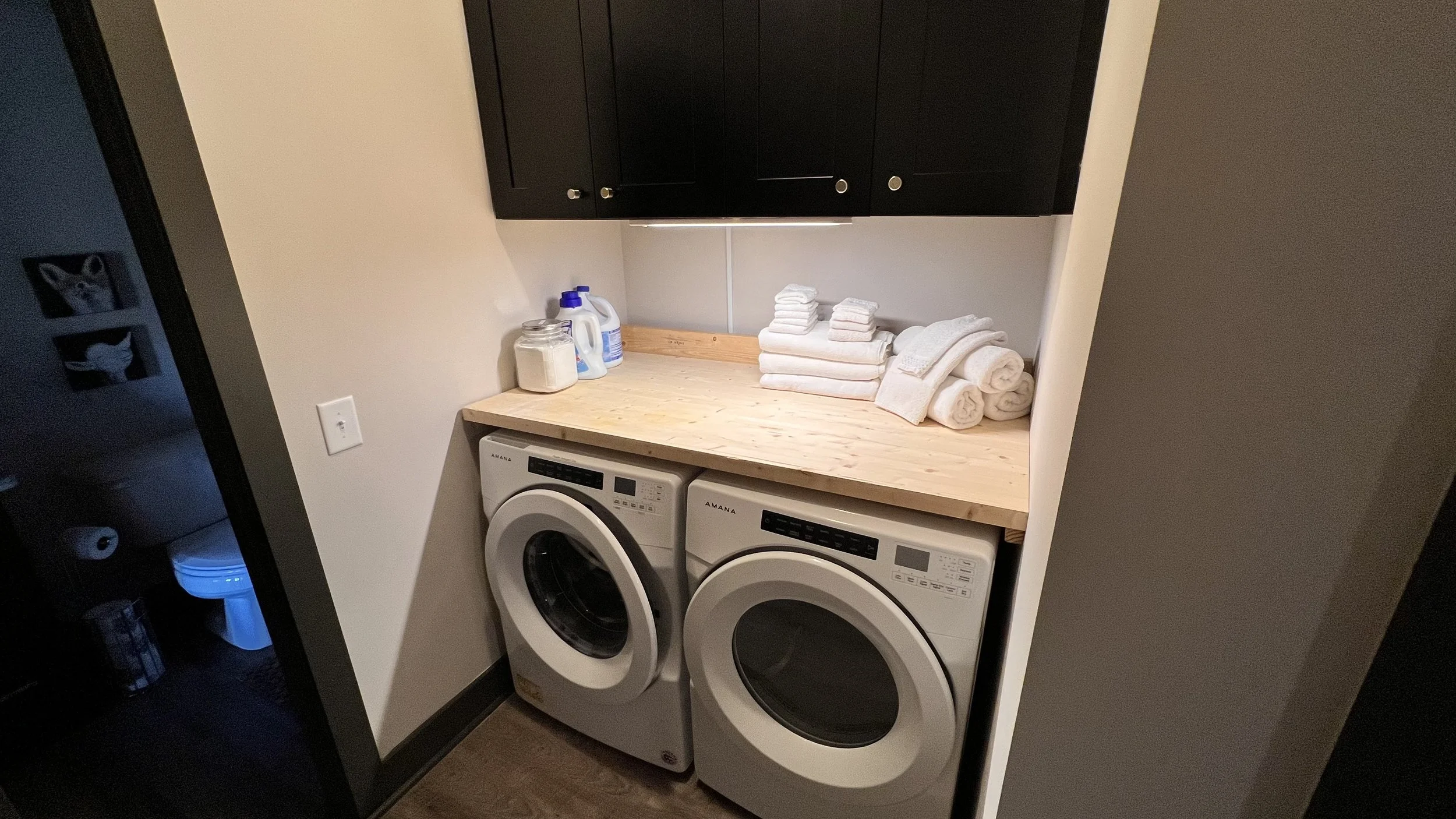 A laundry room with a wooden countertop above two front-loading washing machines. On the countertop are folded white towels, laundry detergent, and a jar of powder.