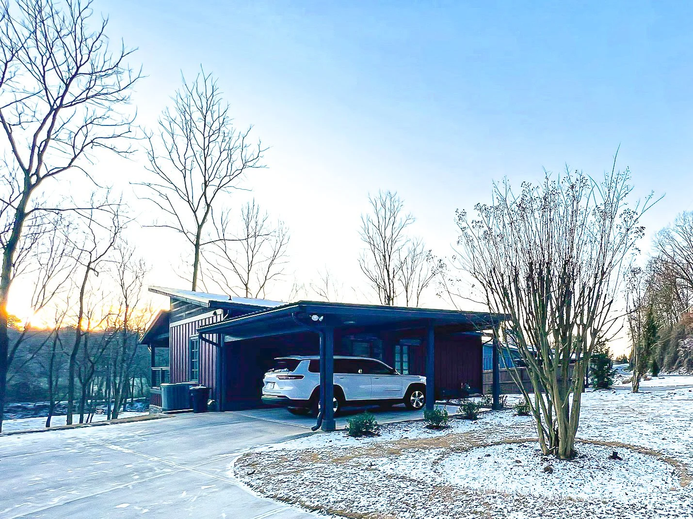 A modern house with a carport, a white SUV parked underneath, and a snow-covered yard with leafless trees and bushes, at sunset or sunrise.