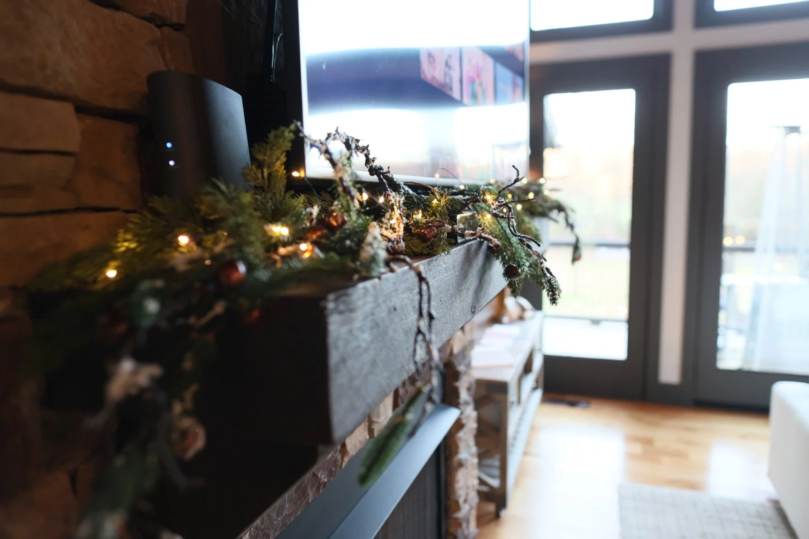 Close-up of a decorated Christmas garland with lights on a fireplace mantel in a living room, with a television and large windows in the background.