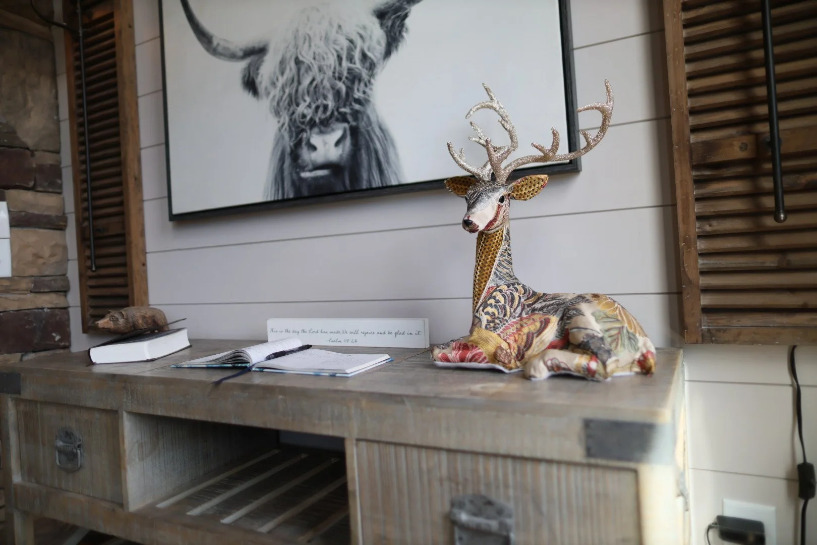 Decorative table with a fabric-covered deer sculpture with antlers, an open book with a pen, a closed book, a small wooden carved animal, a sign with a quote, and a large black-and-white animal portrait hanging on the wall in the background.