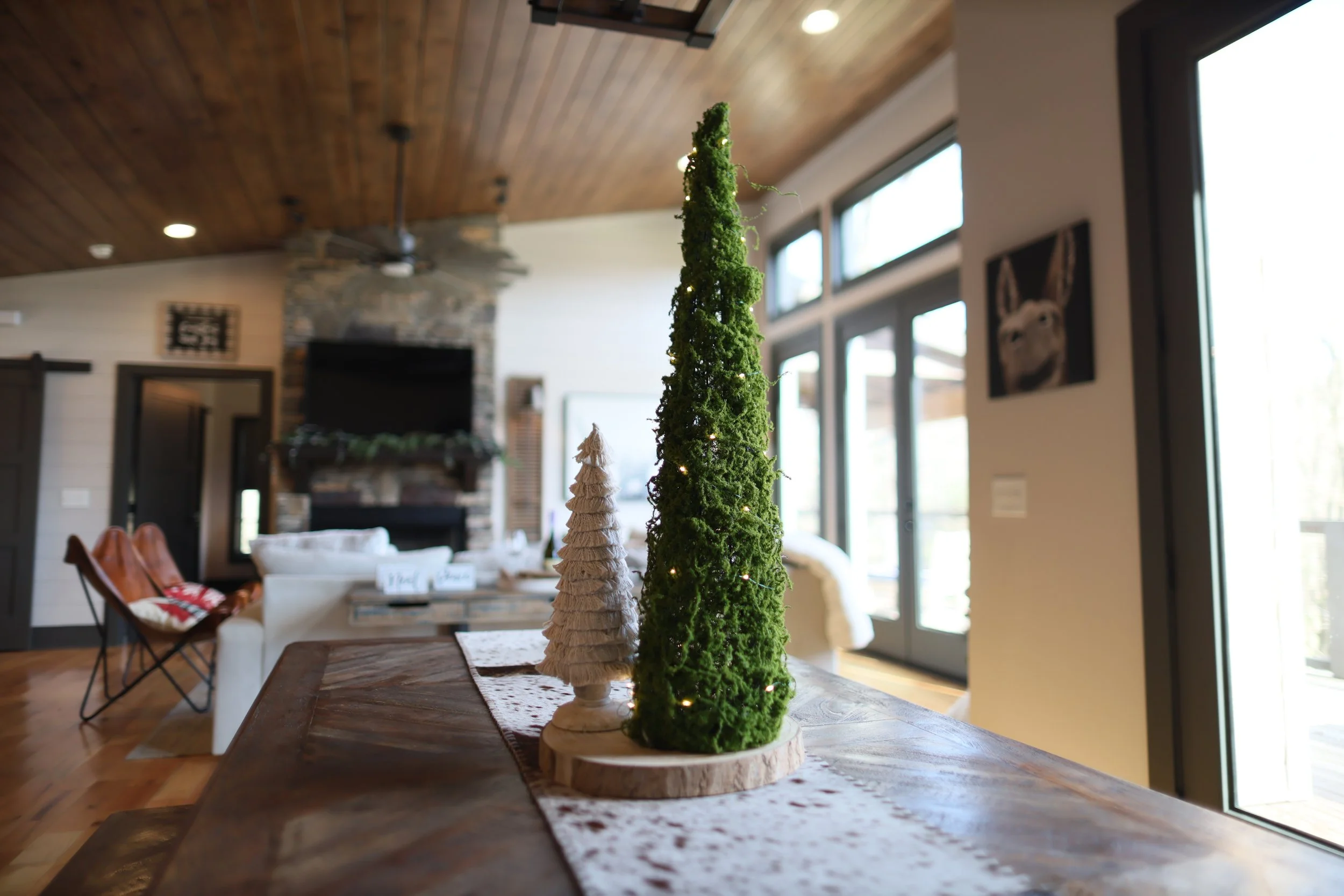 Decorative tabletop Christmas trees, one made of green moss with small lights and another of beige fabric, on a wooden table in a cozy living room.