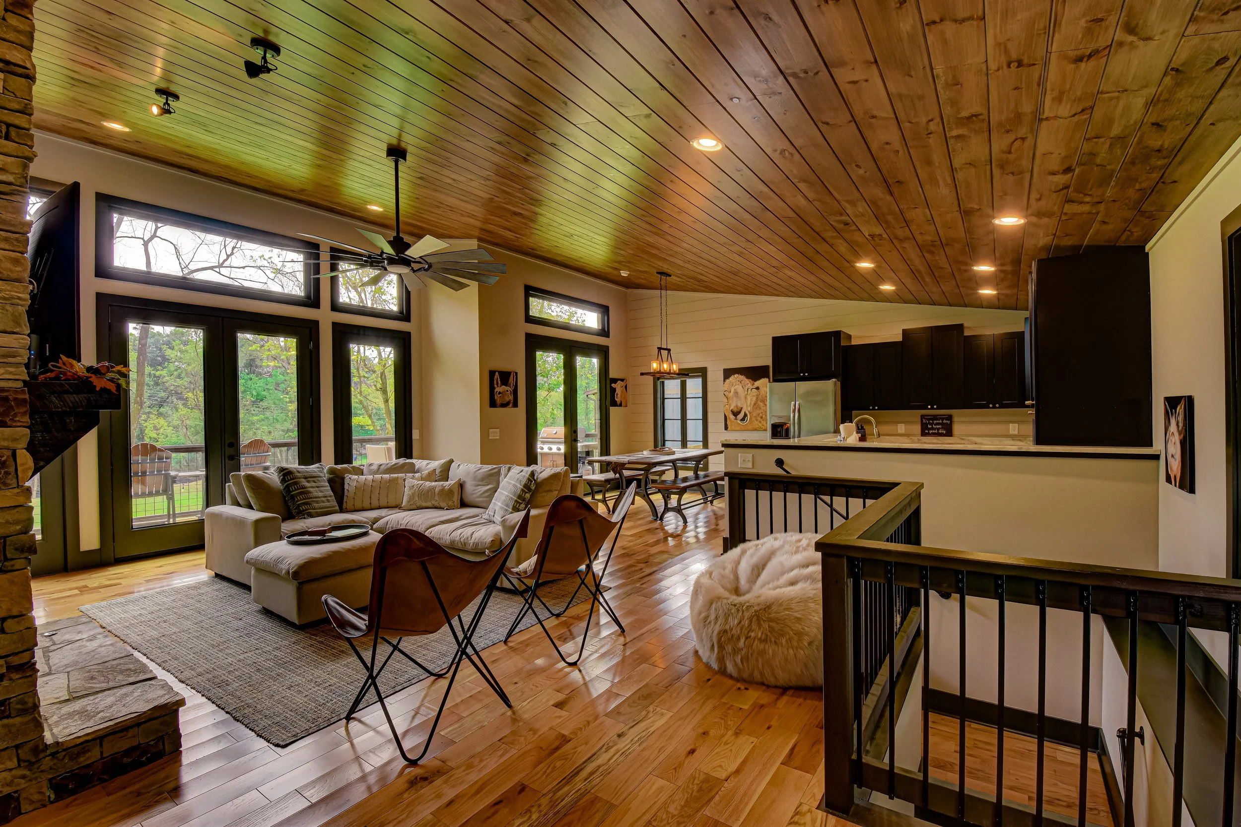 Open-concept living room with large windows, beige sectional sofa, wooden floors, wood-paneled ceiling, black sliding glass doors, and a view of greenery outside.