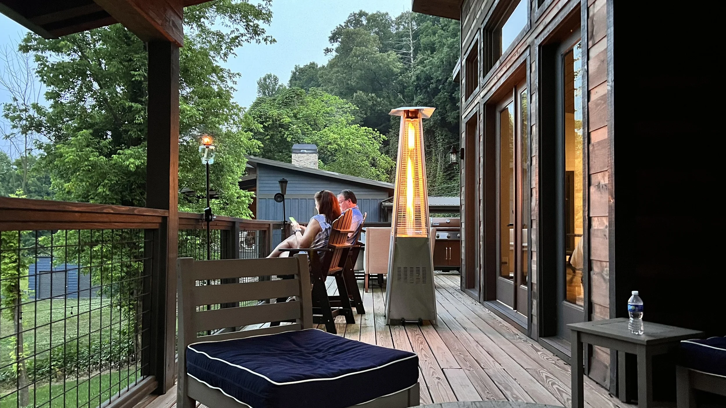 Two people sitting on a balcony, looking at their phones, with a tall patio heater glowing nearby, surrounded by trees and neighboring houses, during dusk.
