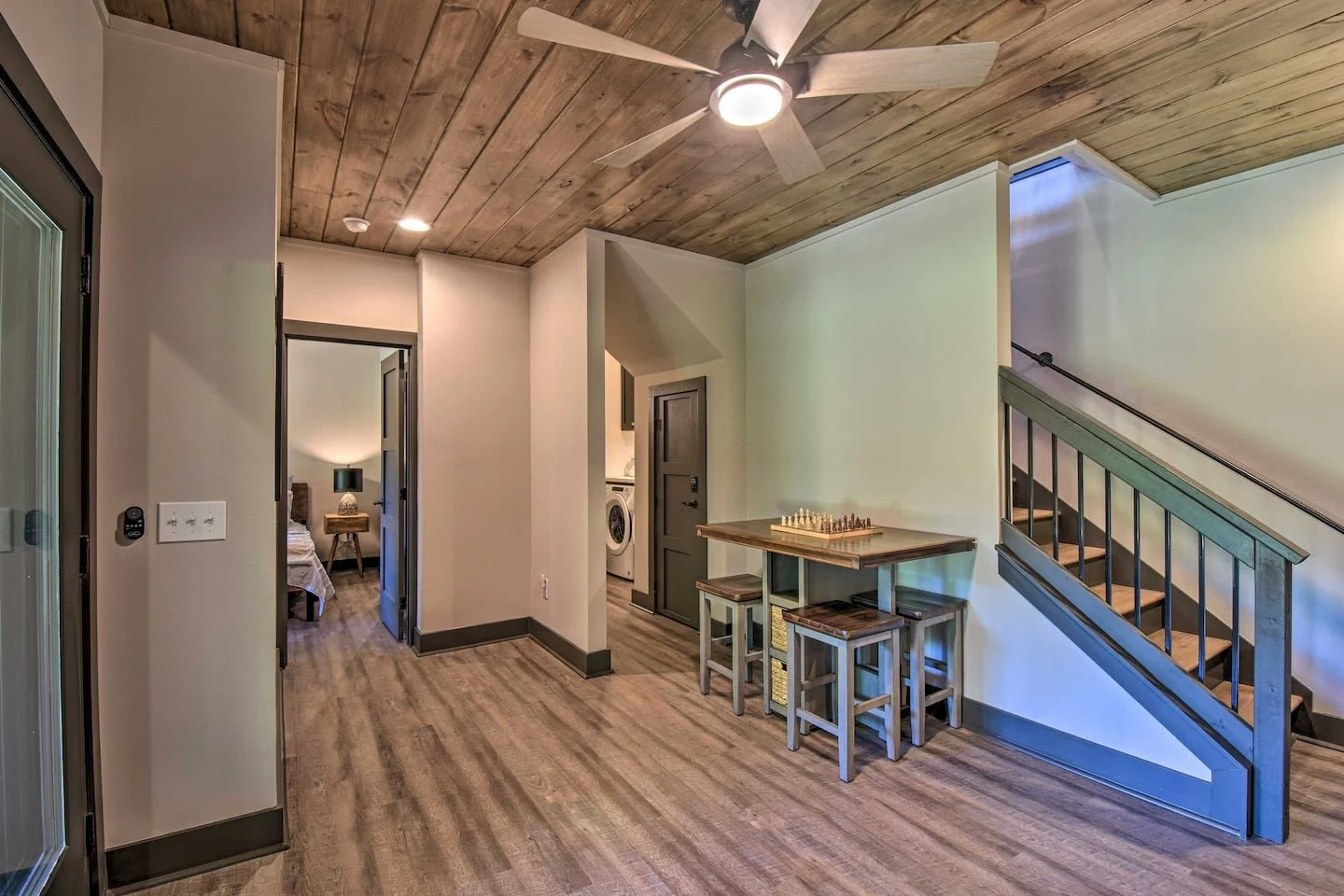 Interior of a home with wooden ceiling, wood flooring, a ceiling fan, a staircase, a small table with a chess set, and a laundry area visible in the background.
