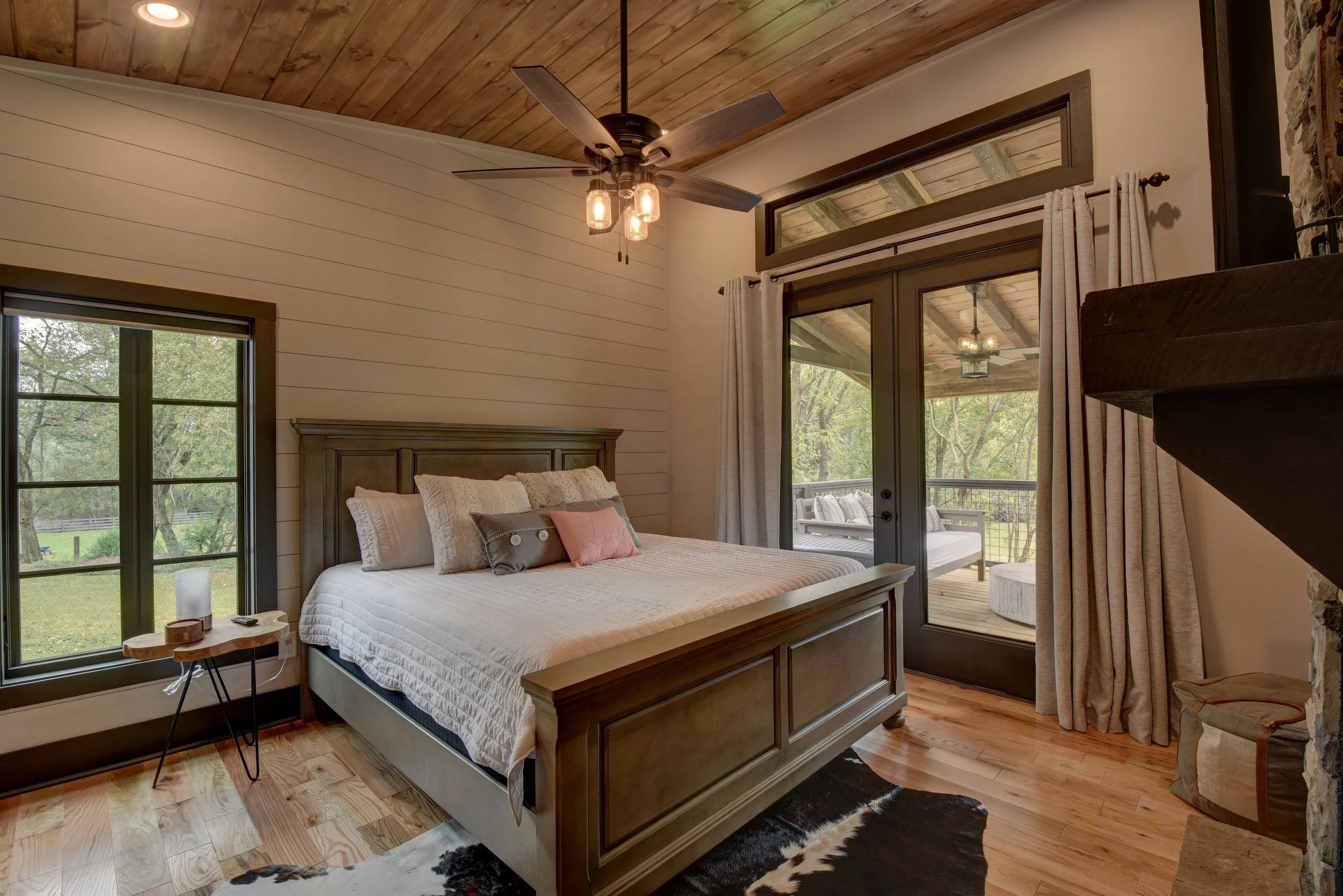 Bedroom with a wood-paneled ceiling, large sliding glass door leading to a porch, wooden bed with pillows, window with black frame, small side table, cowhide rug, and a ceiling fan.