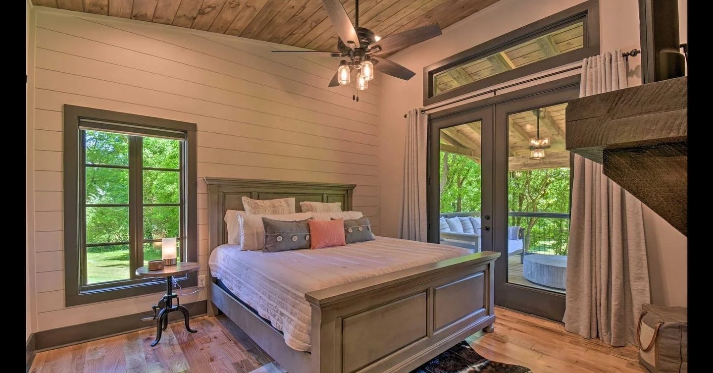 Bedroom with a wooden bed frame, white bedding, multiple pillows, a window showing green trees outside, a bedside table with a lamp, a sliding glass door leading to a balcony with outdoor seating, and a ceiling fan with lights.
