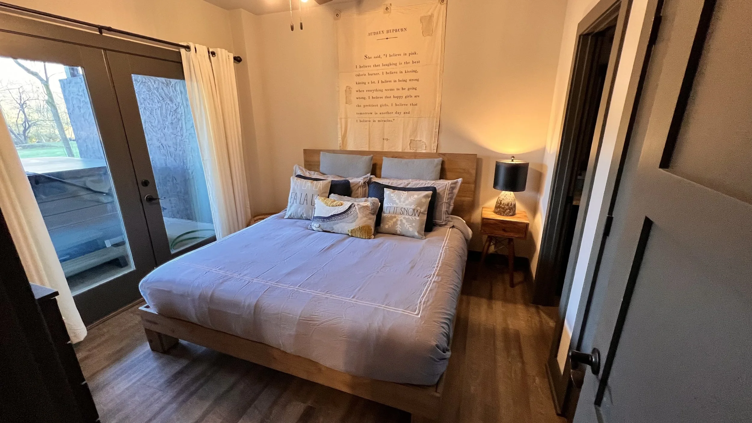 A cozy bedroom with a bed, pillows, and a nightstand with a lamp. There are sliding glass doors with curtains leading to an outdoor area with greenery. A wall decoration features a quote by Audrey Hepburn.