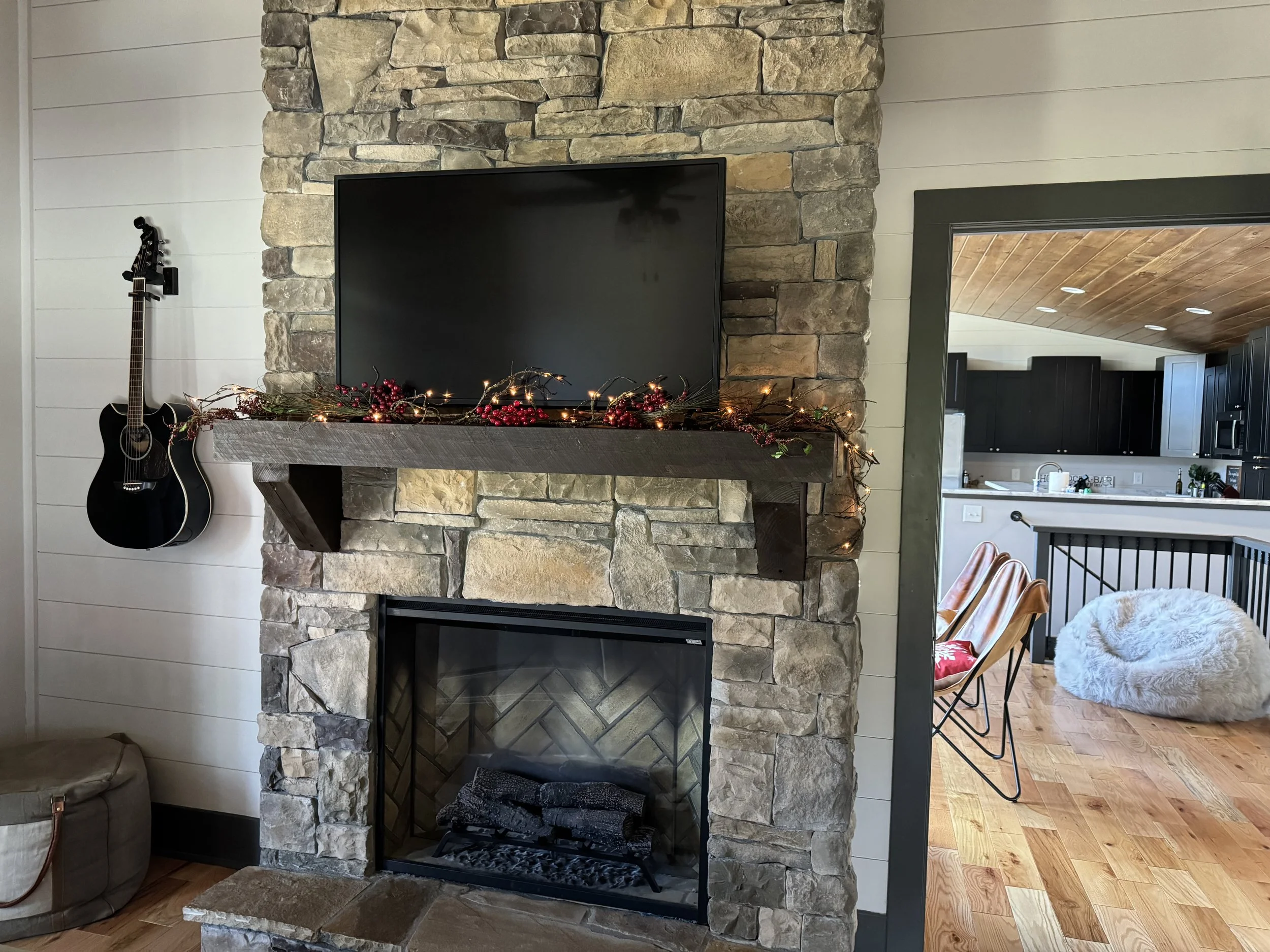 Living room with a stone fireplace, a mounted flat-screen TV decorated with a garland of red berries and lights, a black acoustic guitar hanging on the wall, and visible into the kitchen which has black cabinets, a wooden ceiling, and a cozy seating 