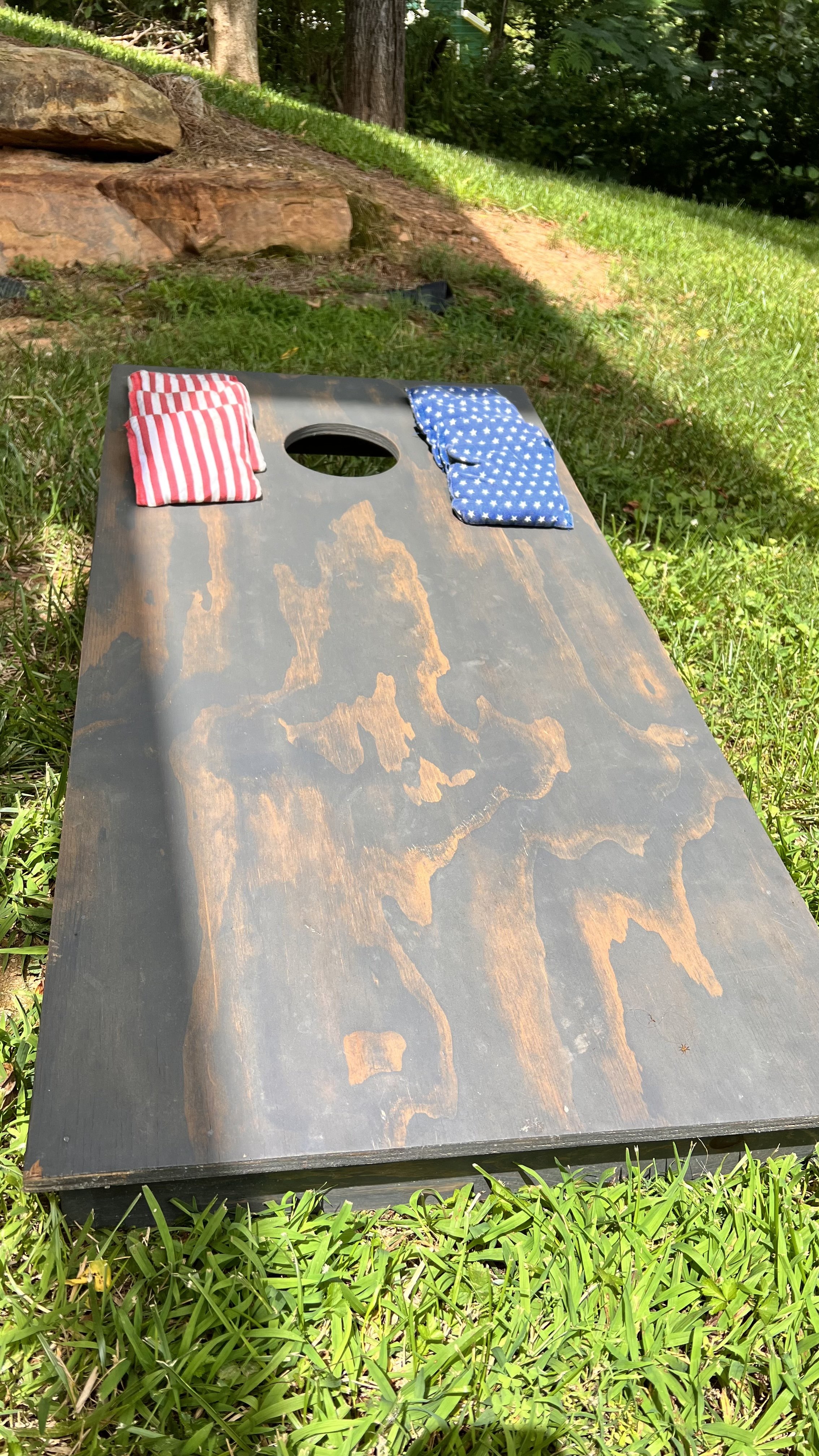 A cornhole game board with red and white striped and blue with white stars cloth bags on top, placed on grass outdoors, with trees and rocks in the background.