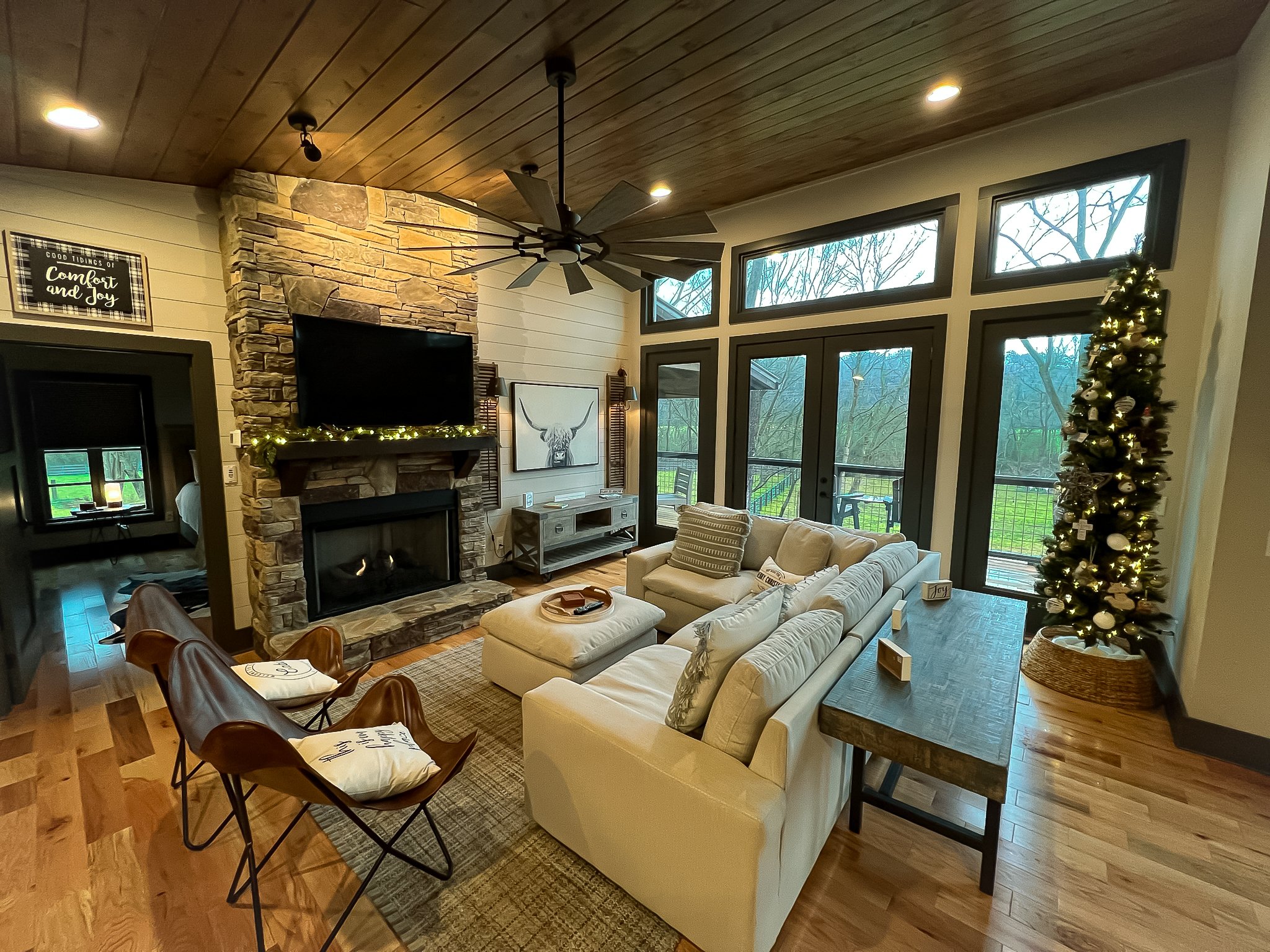 Living room with neutral-colored sectional sofa, two leather chairs, a stone fireplace with a TV, a decorated Christmas tree, large windows, and a wooden ceiling.
