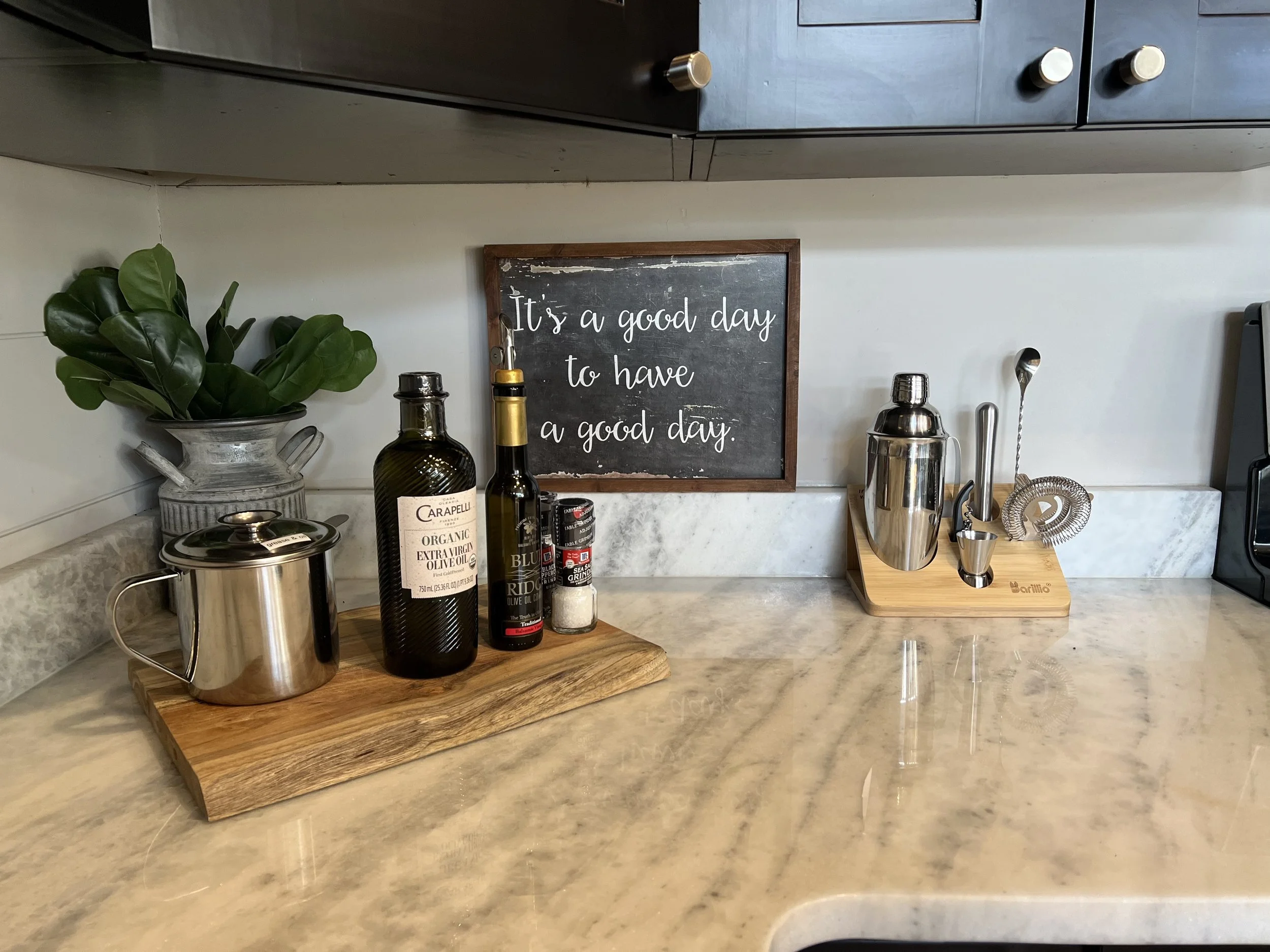 Kitchen countertop with a small chalkboard sign reading 'It's a good day to have a good day,' a metal pot, a bottle of organic extra virgin olive oil, a spice container, a cocktail shaker, and various bar tools arranged on wooden trays, with a plant 