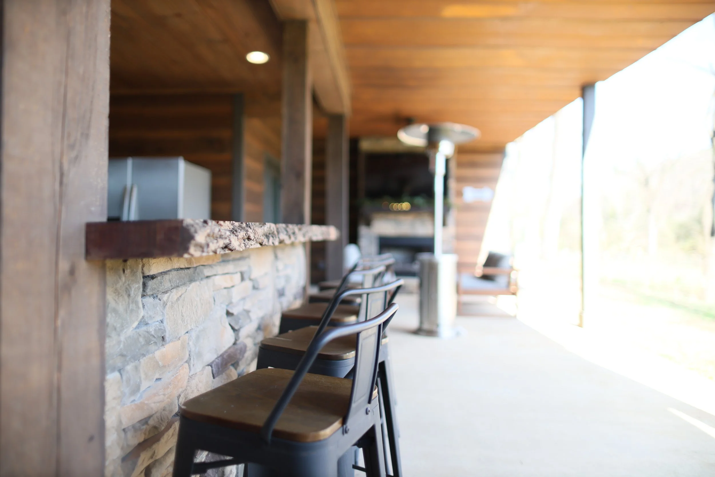 Part of a cozy wooden outdoor patio with a stone bar counter and bar stools, open on one side with a view of bright outdoors.
