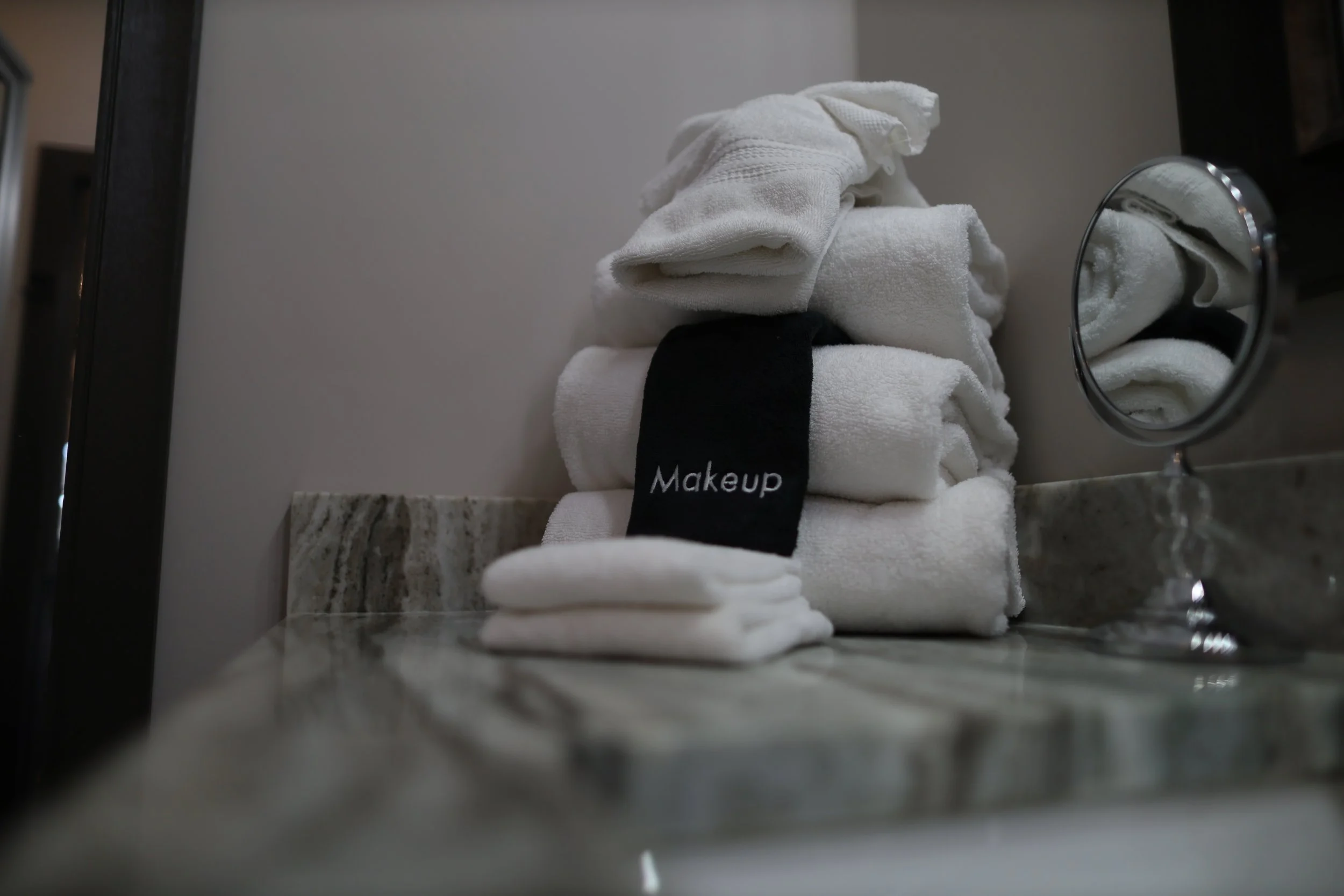 Stack of white towels with one labeled 'Makeup' on a marble countertop next to a small mirror.