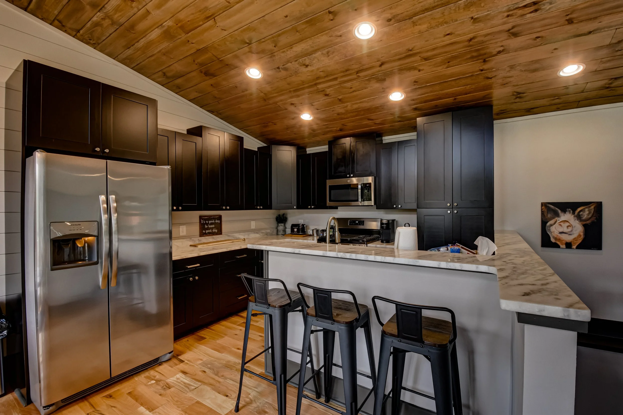 Modern kitchen with dark cabinets, stainless steel refrigerator, marble countertop island with three black barstools, wood ceiling, recessed lighting, and a pig painting on the wall.