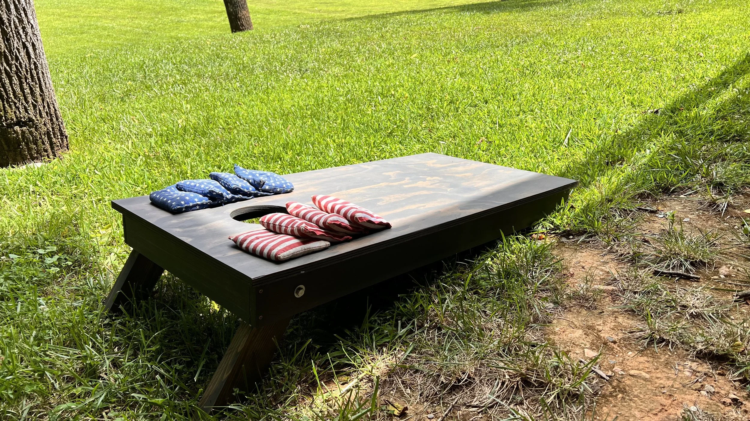 A cornhole game setup outdoors on grass with a black board, four red and white striped bean bags, and four blue bean bags with white stars, on a sunny day near trees.