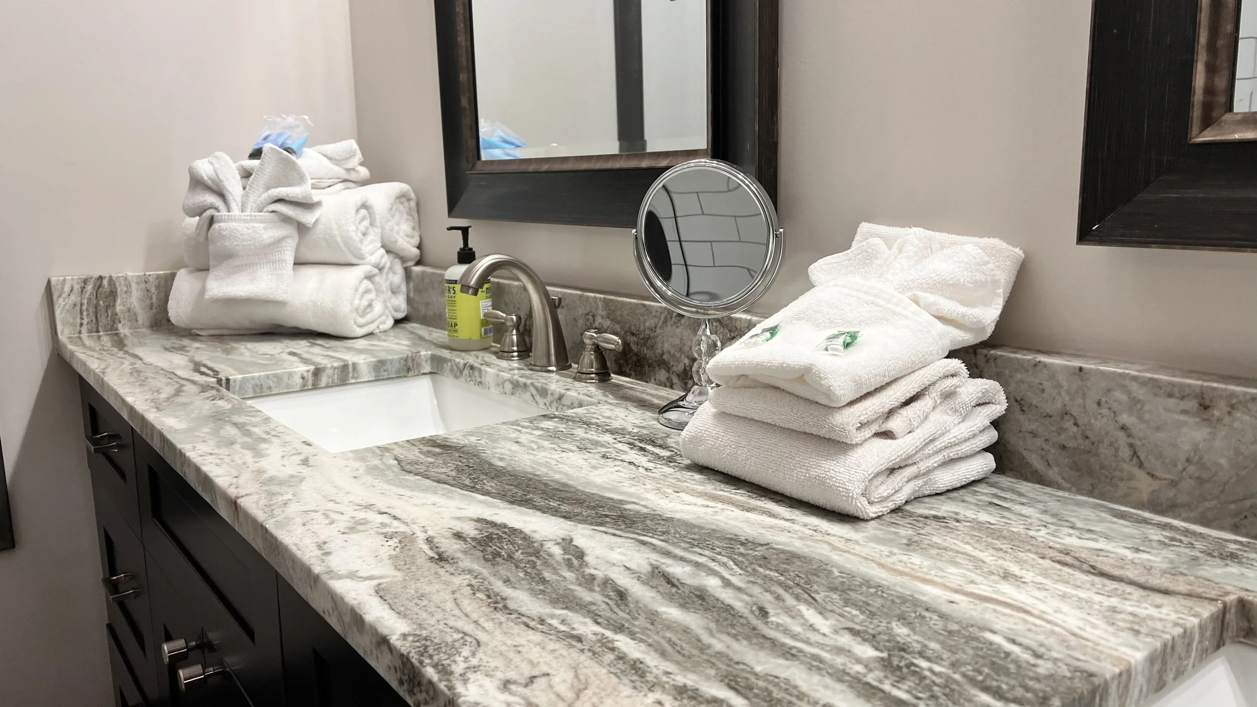 Bathroom countertop with stacked towels, soap dispenser, small round mirror, and two framed mirrors on the wall.