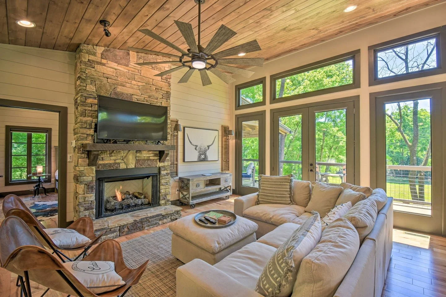 Living room with large beige sectional sofa, stone fireplace, and a ceiling fan. Large windows and glass door show greenery outside.