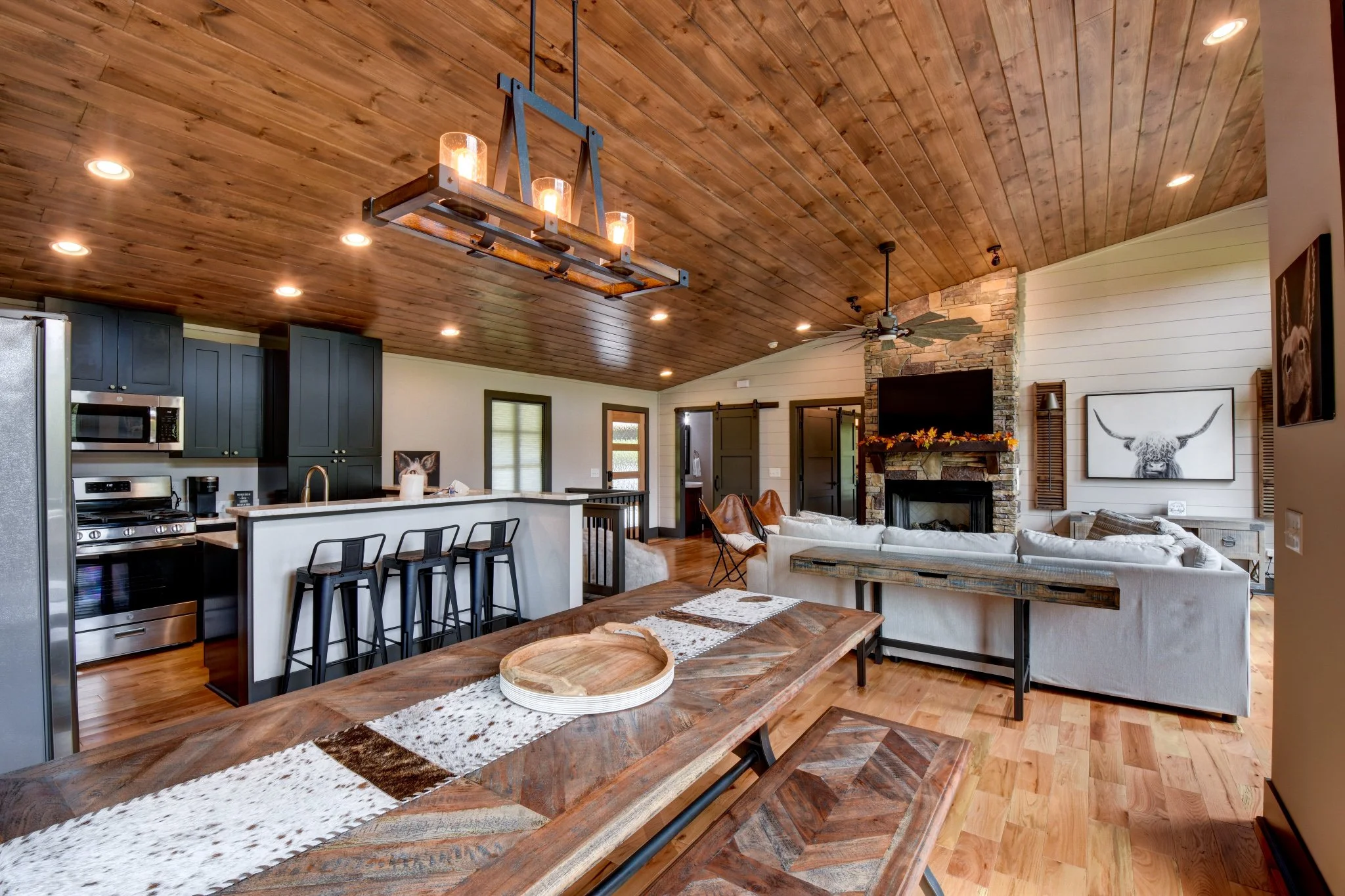 Open-concept living room and kitchen with wooden ceiling and flooring, white walls, a stone fireplace with a TV, a white sectional sofa, and a wooden dining table with benches.