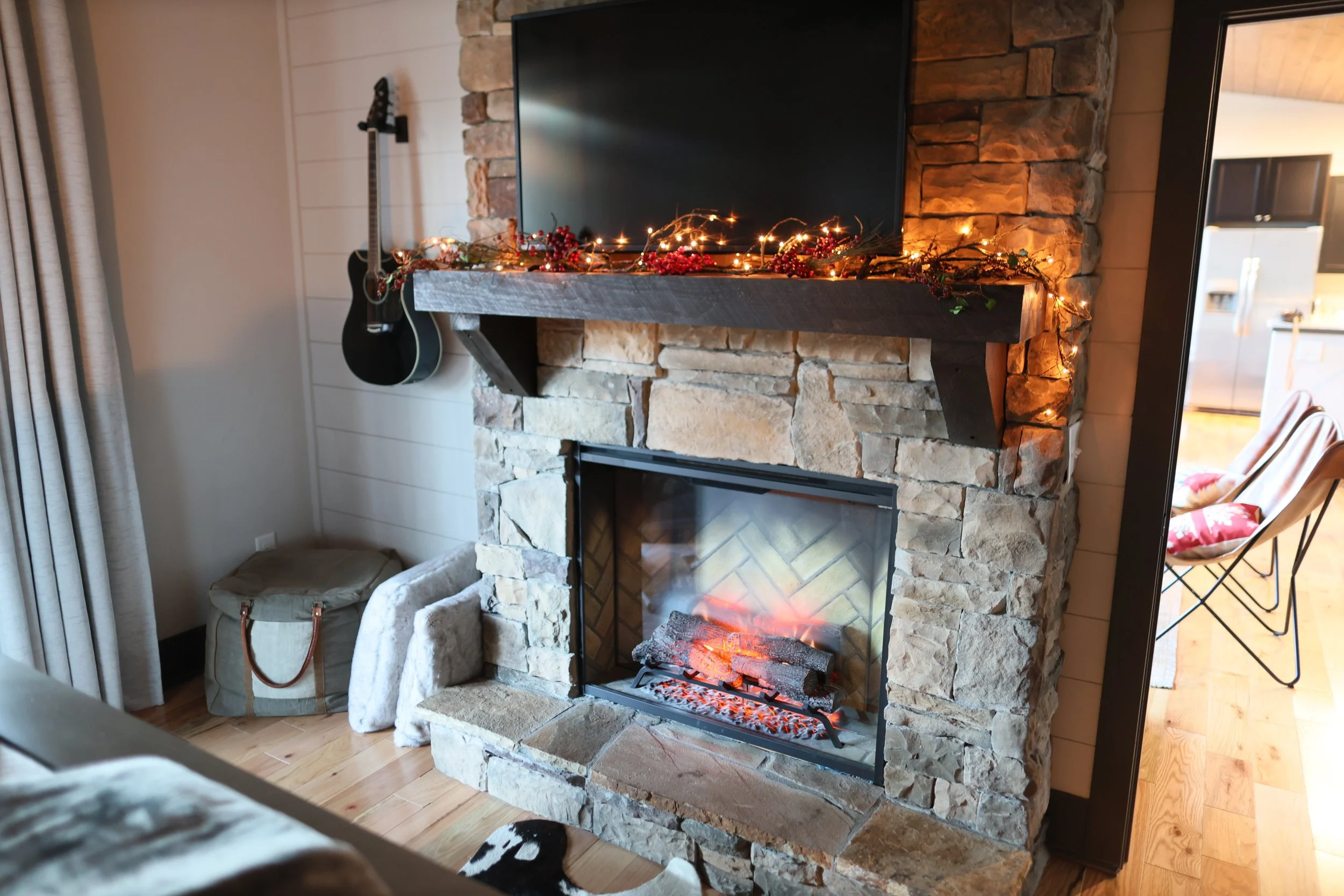 Living room fireplace with a stone surround and a dark wooden mantel decorated with Christmas garlands and string lights, with a flat-screen TV mounted above.