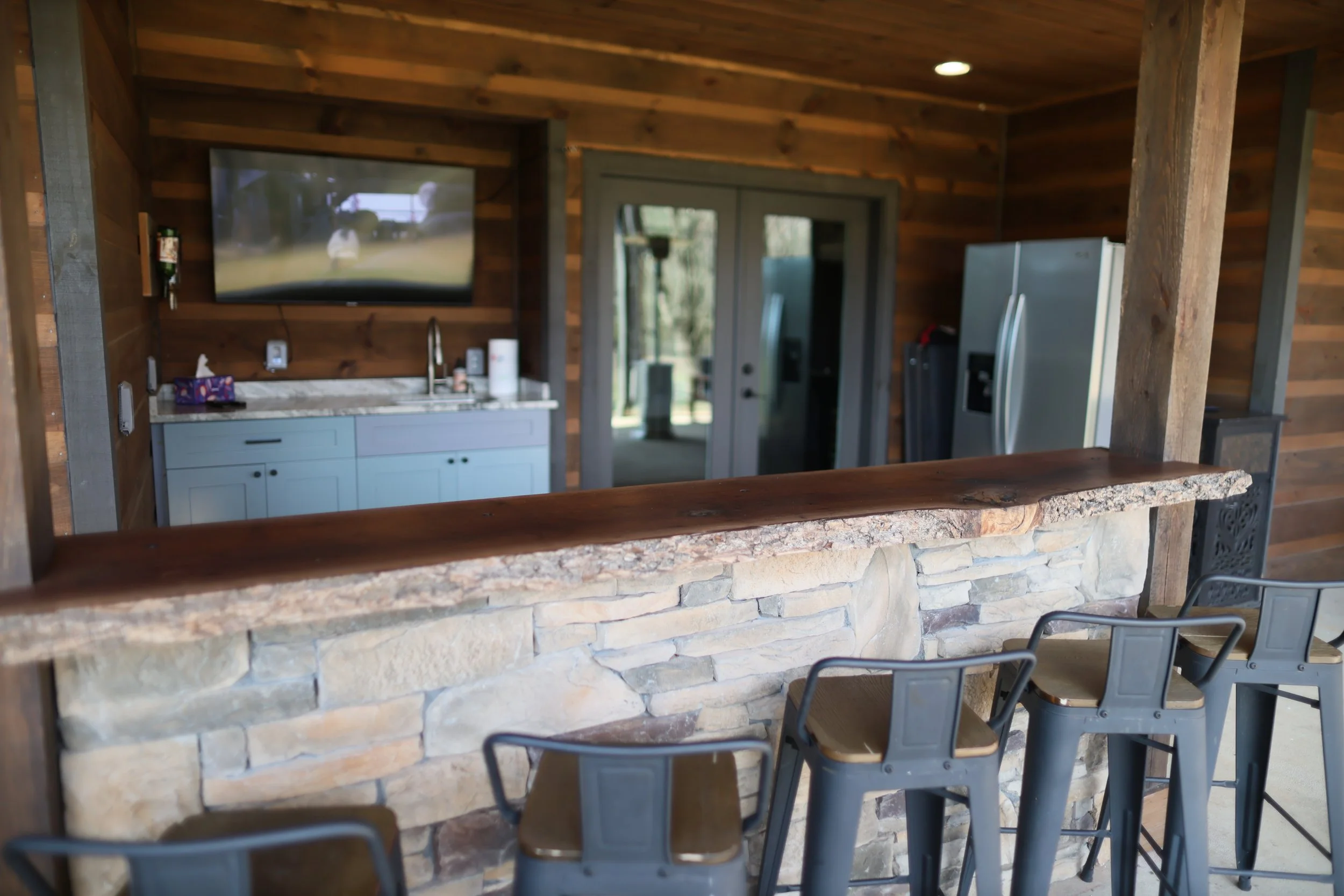 Outdoor bar area with four metal and wood chairs in front, stone and wood counter, wooden walls, and a TV above a small sink, with a sliding glass door leading outside.