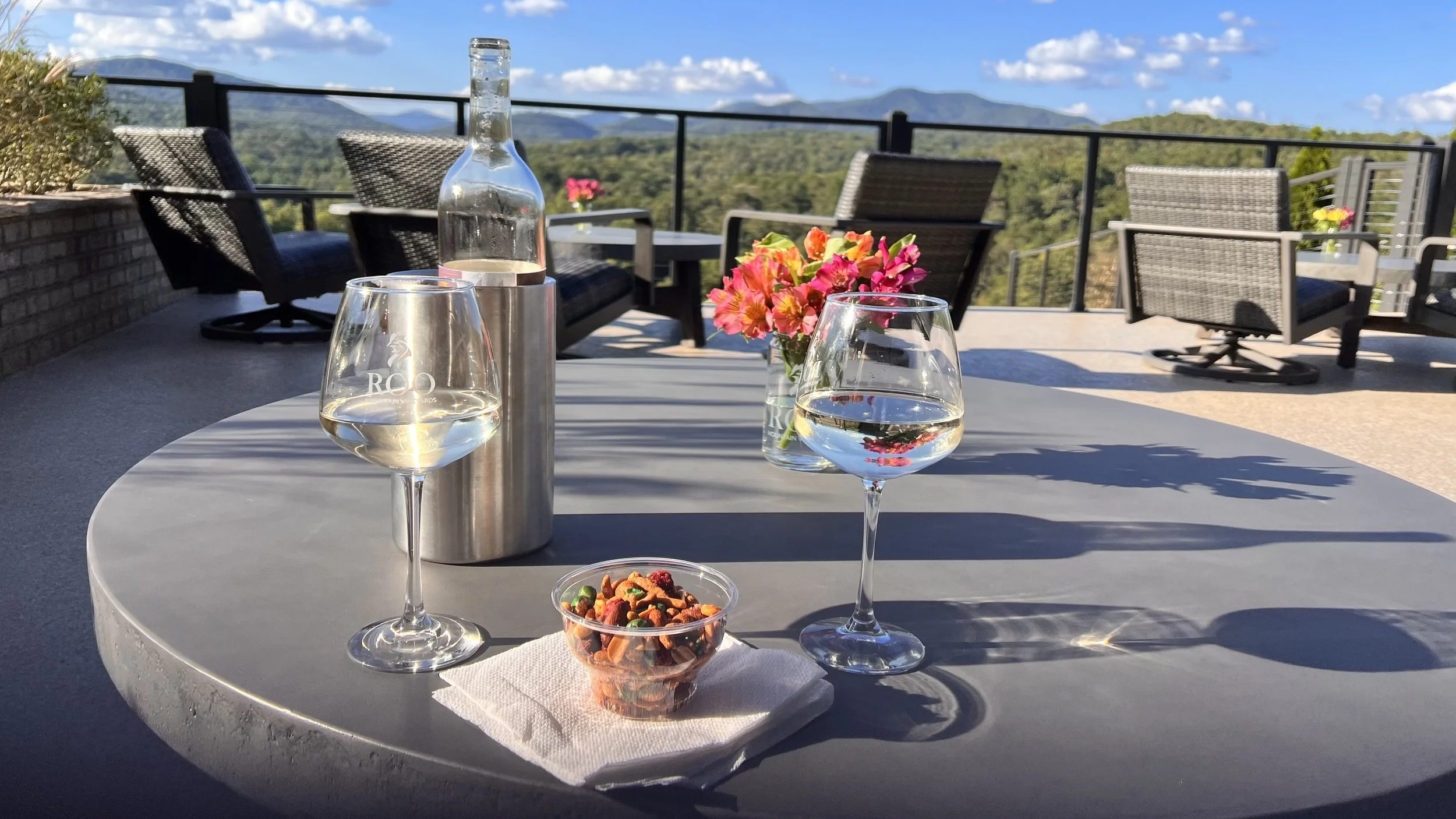 Outdoor patio table with two glasses of white wine, a water bottle in a metal holder, a small bowl of trail mix, and a vase with pink and orange flowers, overlooking a scenic mountain view under a blue sky.