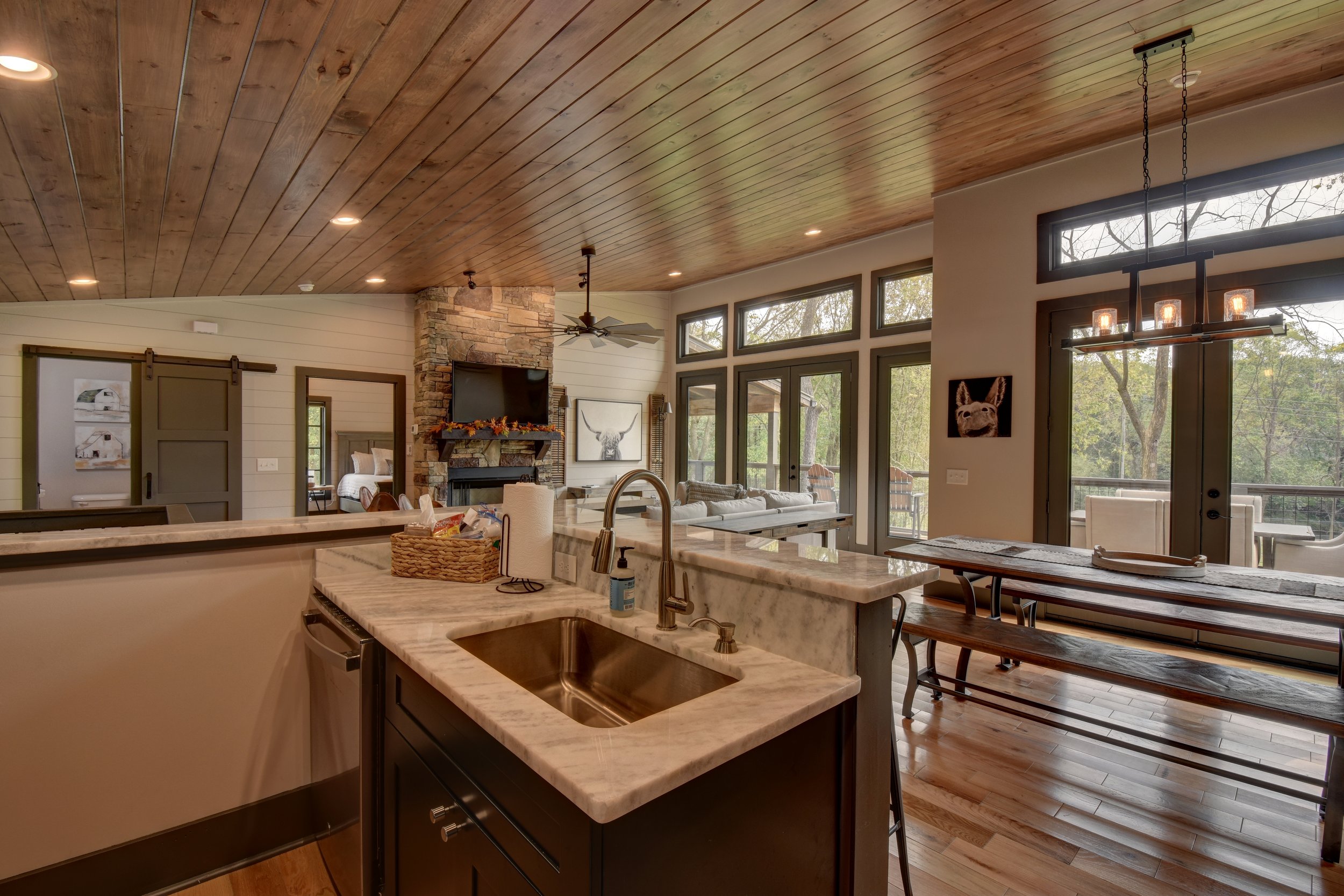 Open-concept living area with a wooden ceiling, stone fireplace, and large windows showing trees outside. A kitchen island with a sink and soap dispenser, a wooden dining table with benches, and a lounge area with a sofa and artwork of a dog.