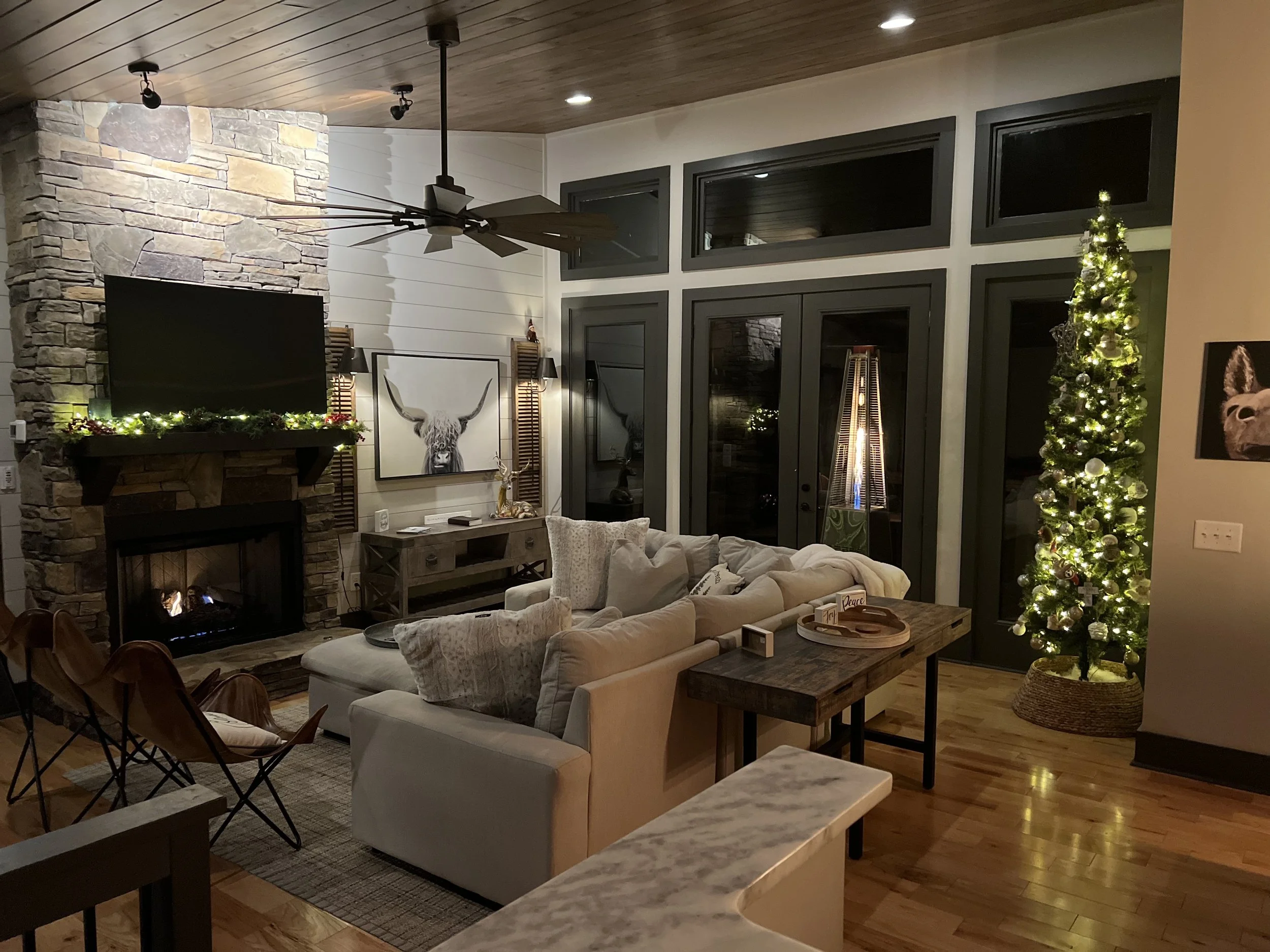 Living room decorated for Christmas with a lit Christmas tree, a stone fireplace with a TV, a ceiling fan, and a sectional sofa with cushions.