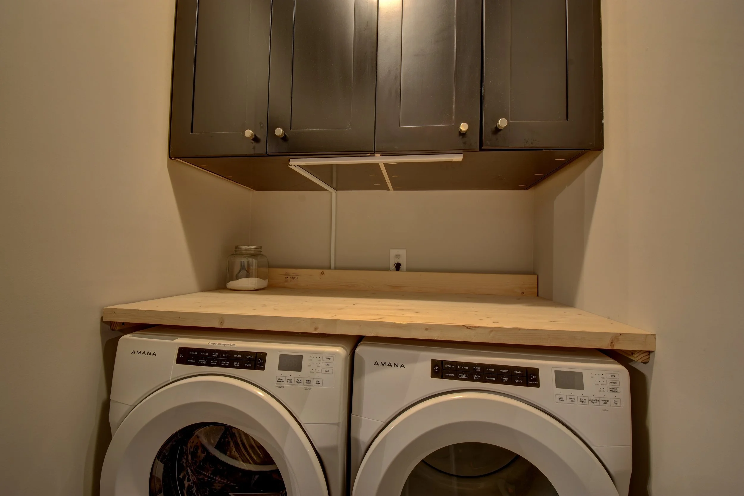 A laundry room with a wooden countertop above two front-loading washing machines, a glass jar on the countertop, and dark wall cabinets above.