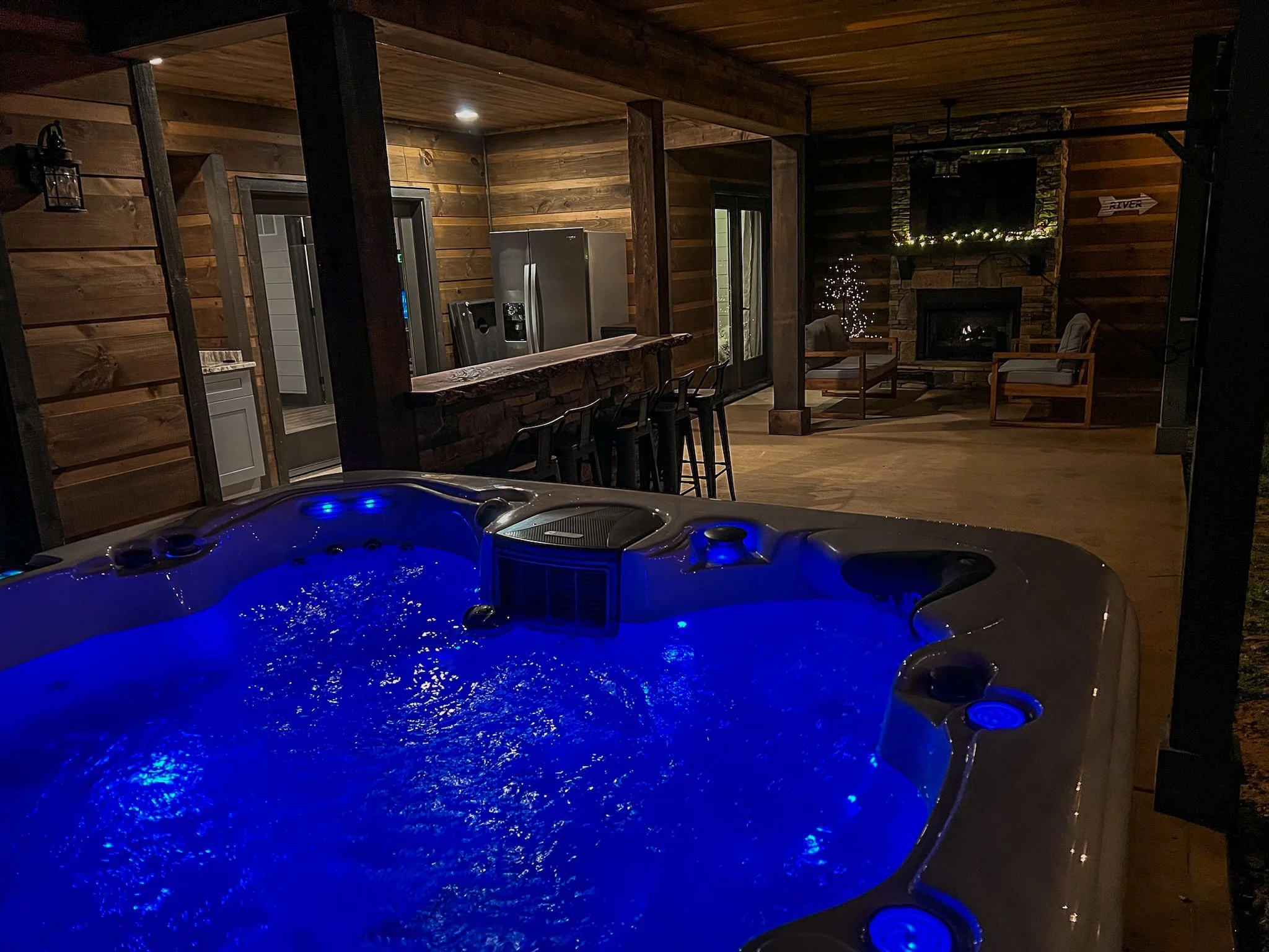 A backyard outdoor living space at night featuring a lit hot tub with blue lights in the foreground, a wooden deck with barstools, a fireplace with a TV above, surrounded by wooden walls and furniture.