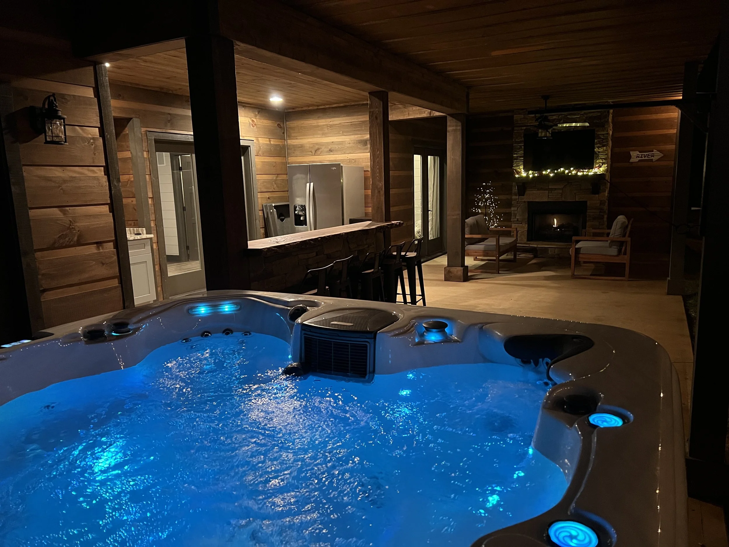 Indoor hot tub with blue LED lights in a cozy cabin living room with wood-paneled walls, fireplace, and seating area.