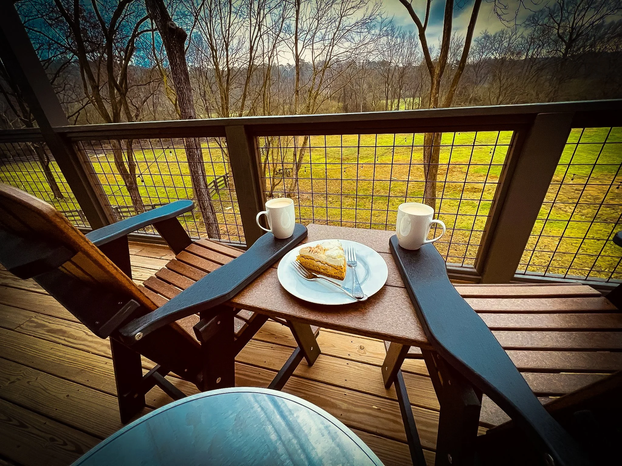 On a wooden balcony, a small table with a piece of pie on a white plate, two white mugs, and a fork and knife. There are two chairs, one of which is visible, with a view of a grassy field and leafless trees in the background.