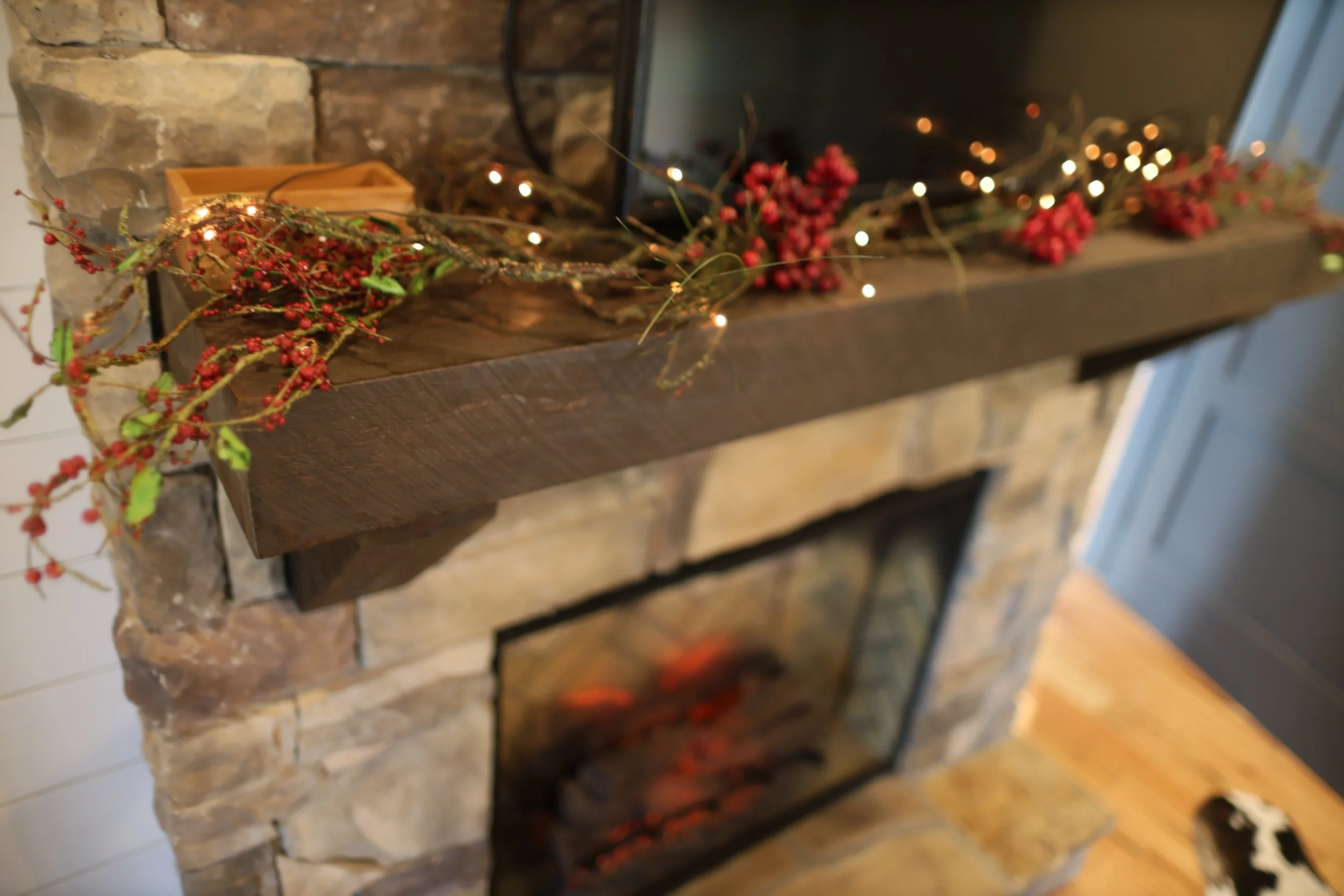 Decorated fireplace mantel with berry garland and fairy lights over a rustic stone and wood fireplace.