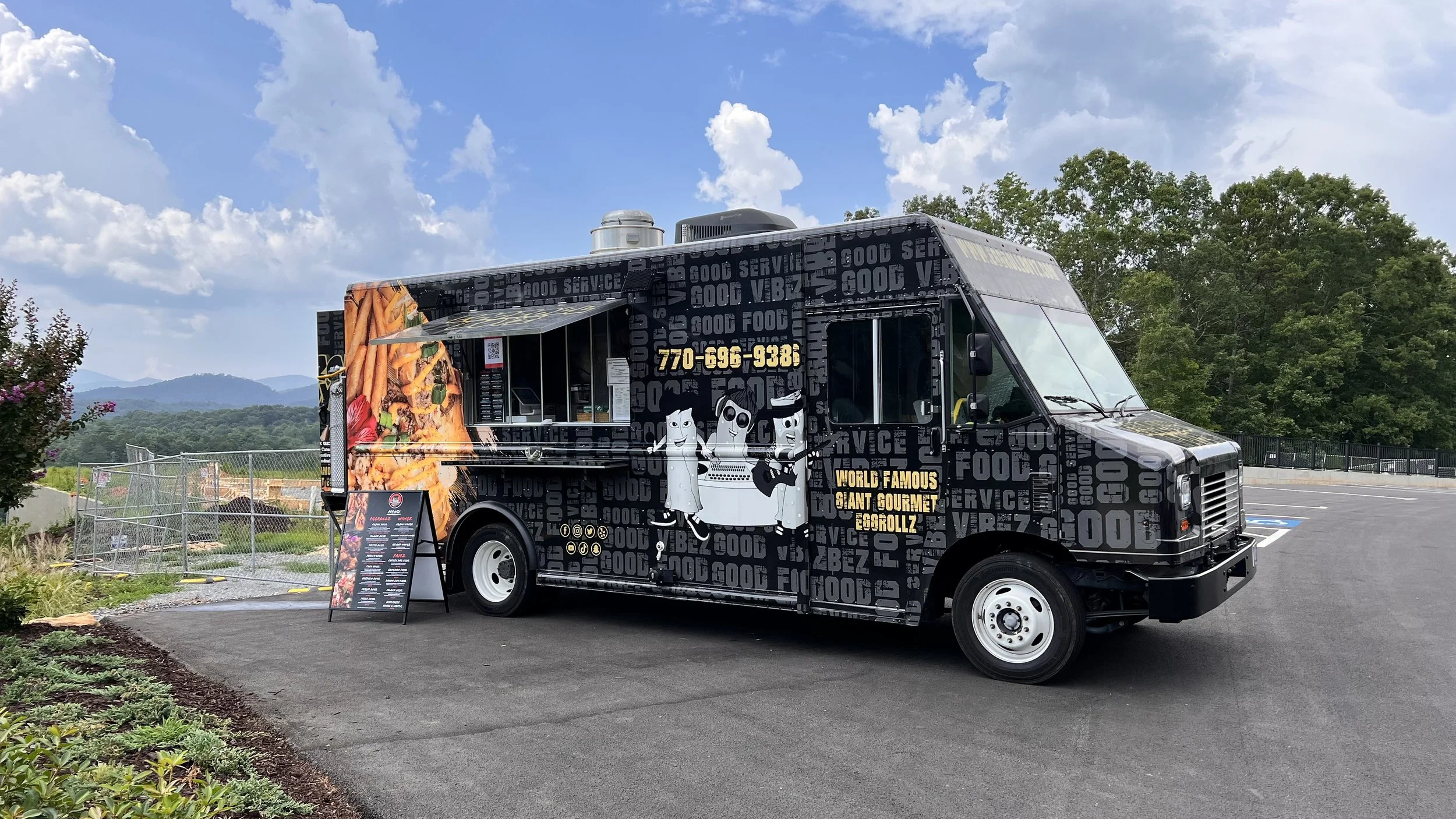 Black food truck with cartoon characters and text, parked on asphalt with a menu sign outside. Background features greenery, trees, and mountains under a partly cloudy sky.
