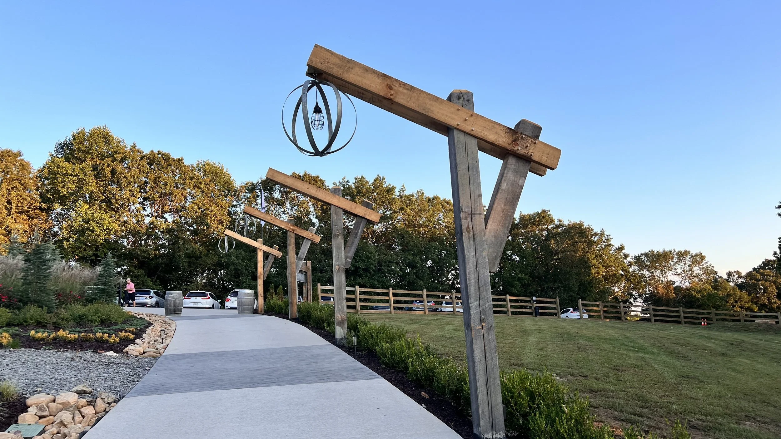 Outdoor setting with a paved walking path, decorative wooden beams with metal rings and hanging lanterns, surrounded by green grass, trees, and parked cars in the background.