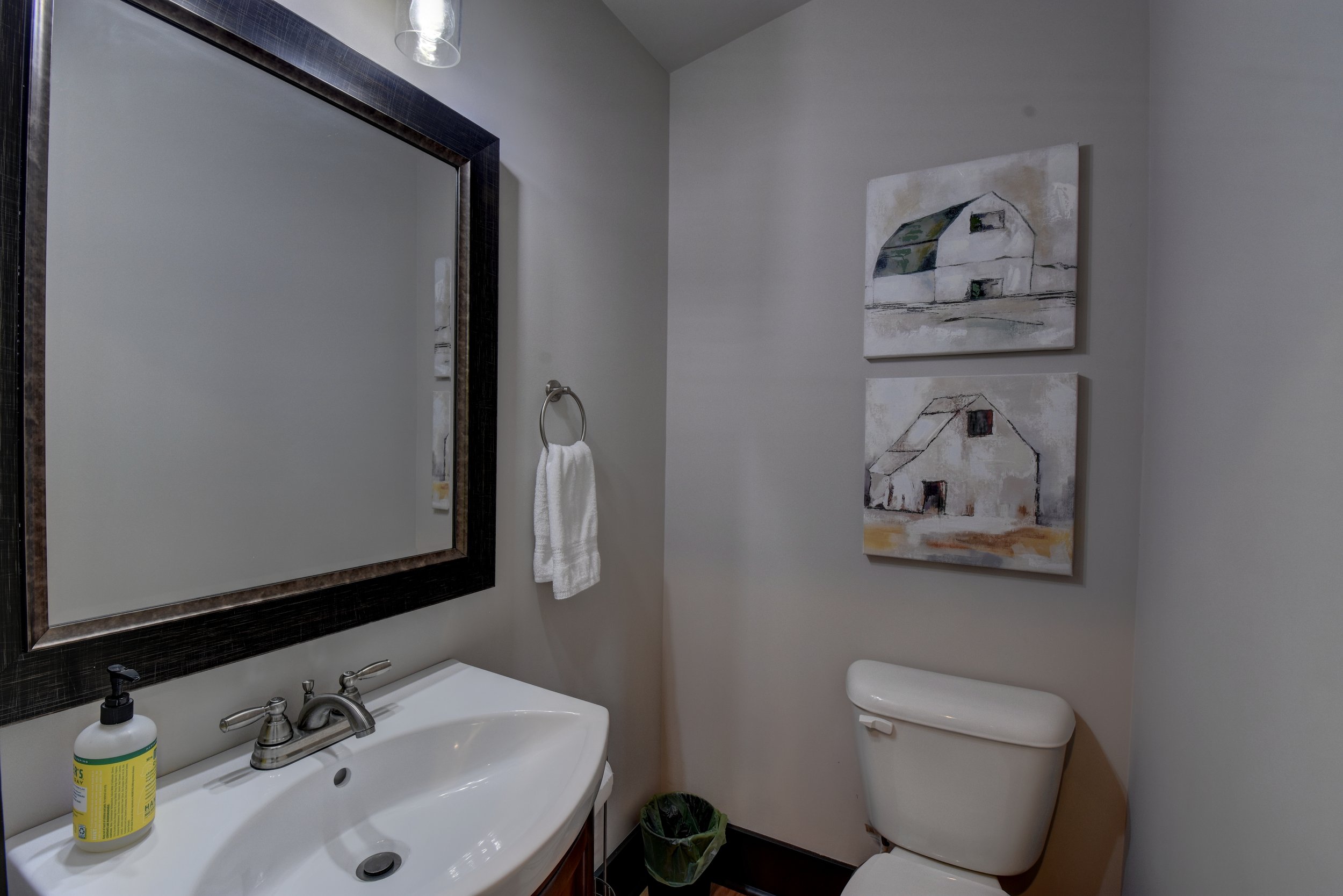 A bathroom with a large rectangular mirror, a white sink with a soap dispenser, a towel ring with a white towel, a toilet, two paintings of barns on the wall, and a small trash can.