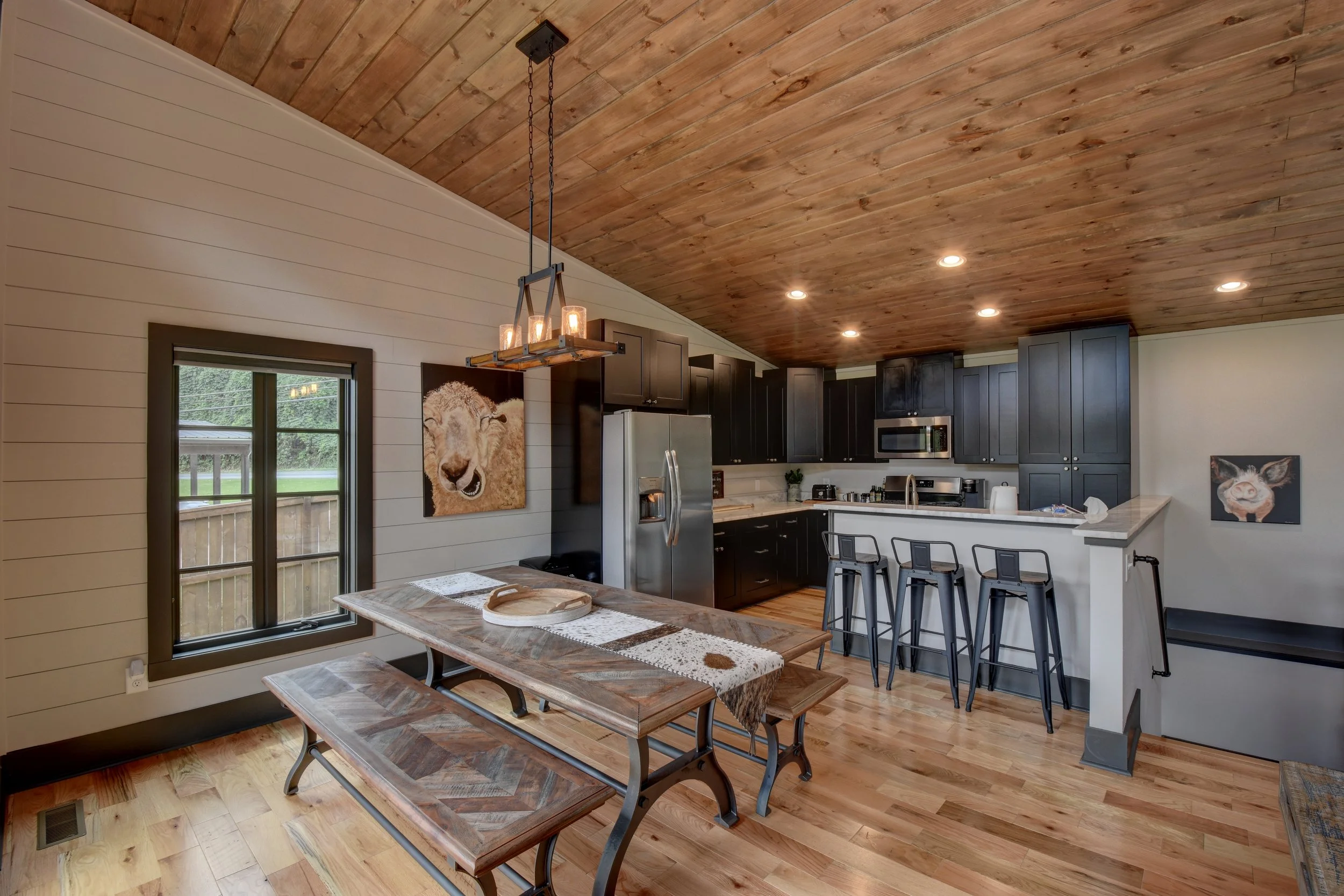 A kitchen with dark cabinets, stainless steel appliances, and a wood-paneled ceiling. There is a dining area with a wooden table and bench, a window, and artwork on the walls.