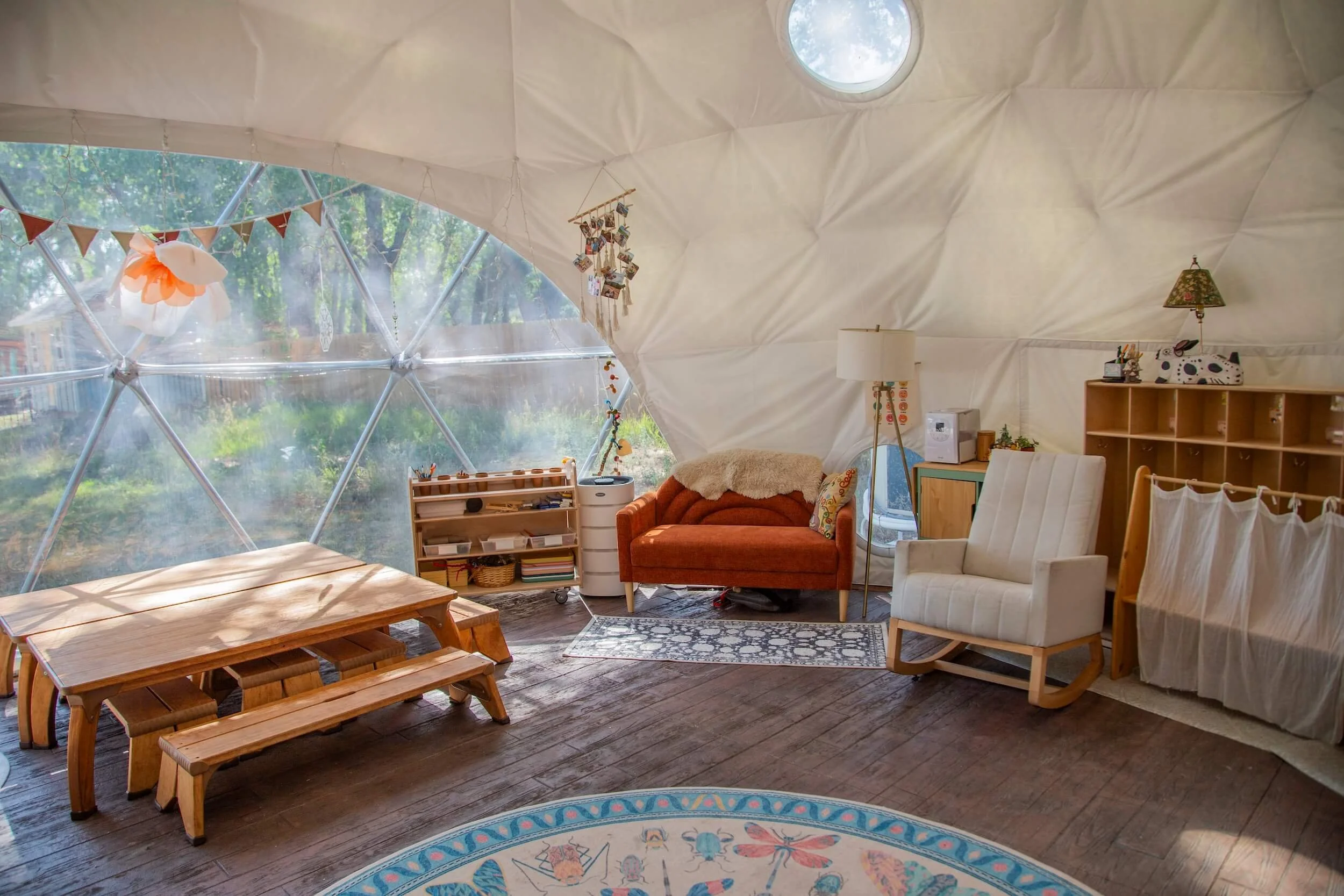 Interior of a cozy dome-shaped room with large windows showing trees outside. The room has wooden furniture, including a bench, a white armchair, and a wooden bookshelf. Decorations include a hanging photo display, a lamp with a floral shade, and bun