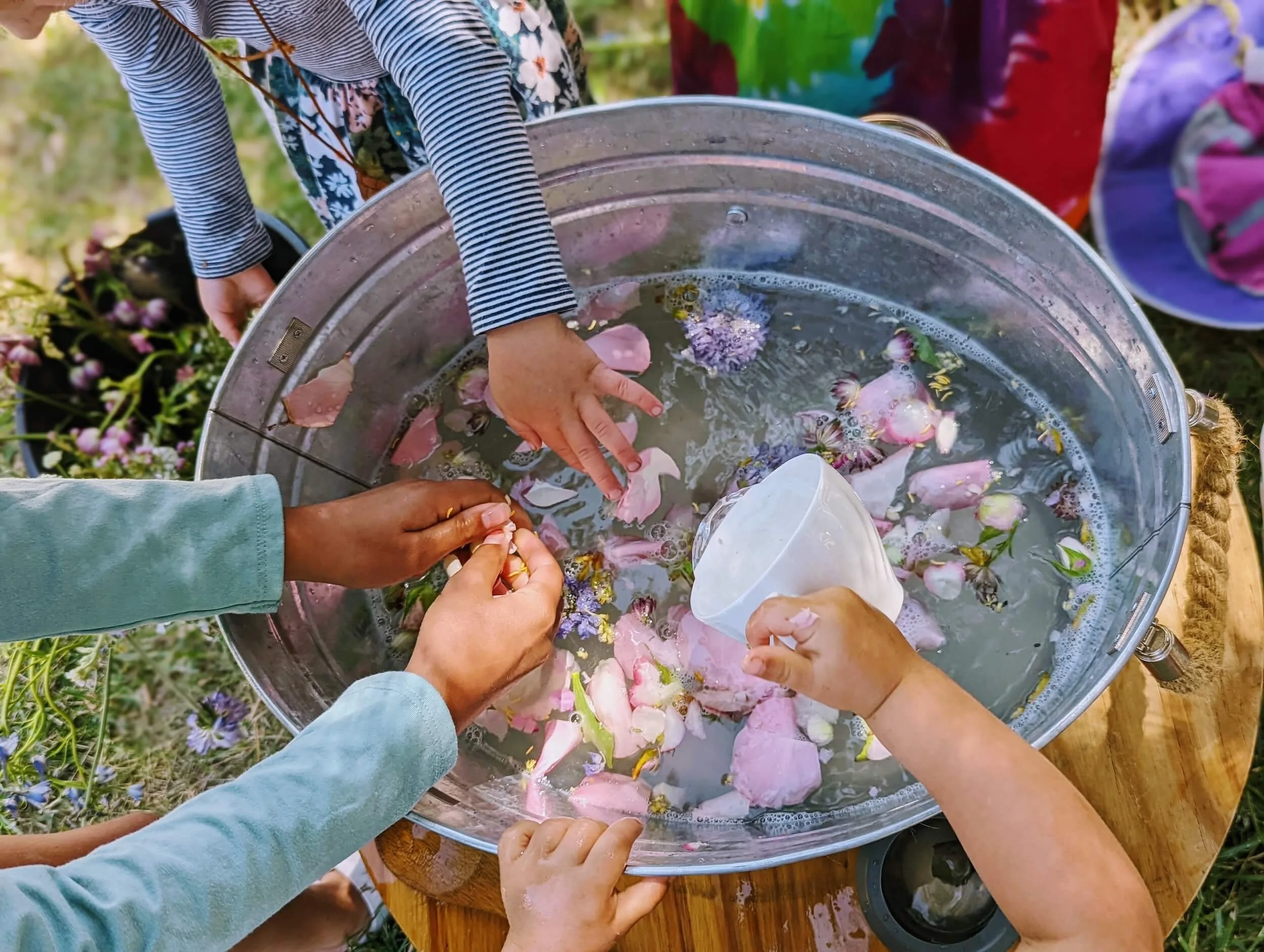 People gathered around a large metal basin filled with water and flower petals, participating in an activity outdoors.