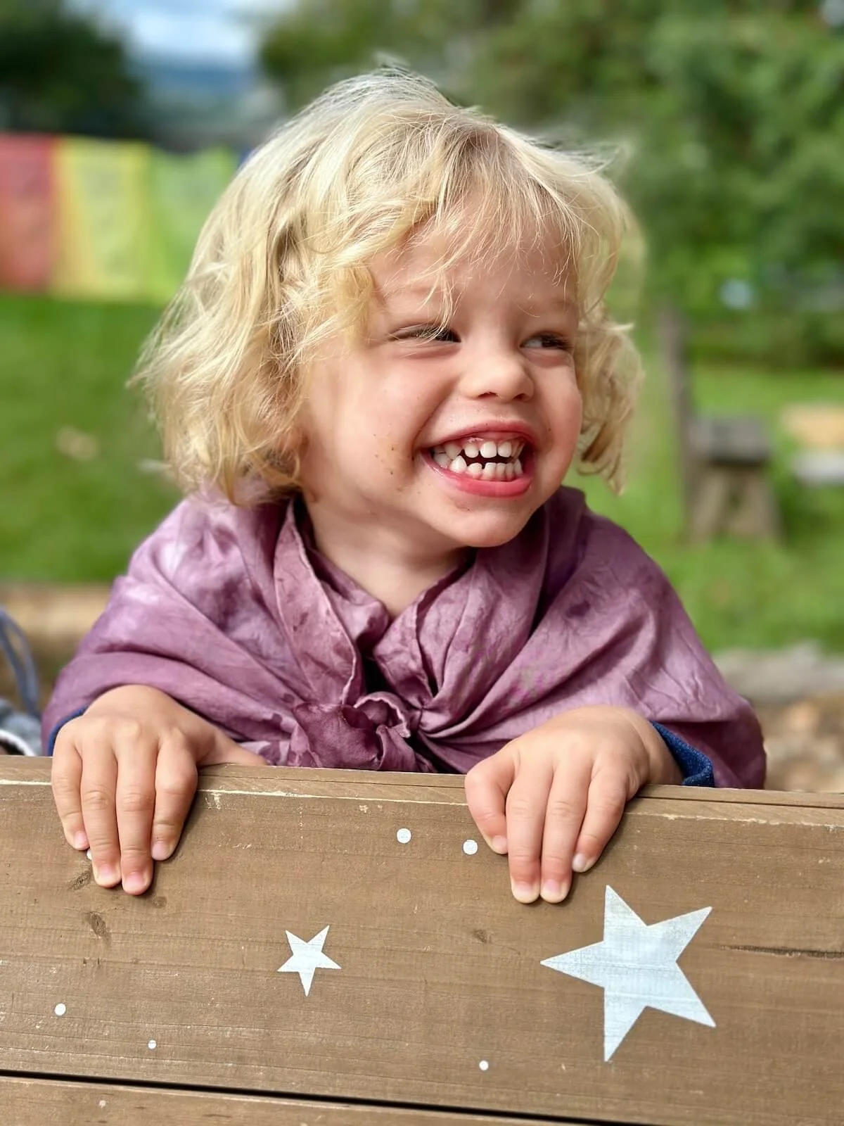 A young girl with curly blonde hair smiling and showing missing front teeth, leaning on a wooden surface with star decorations, outdoors with green trees and grass in the background.