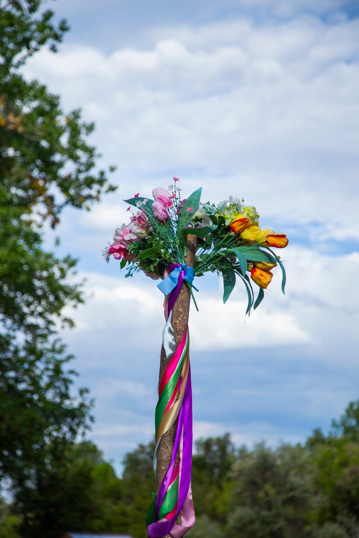 Colorful bouquet of flowers tied to a stick with multicolored ribbons, held up in an outdoor setting with trees and a cloudy sky in the background.